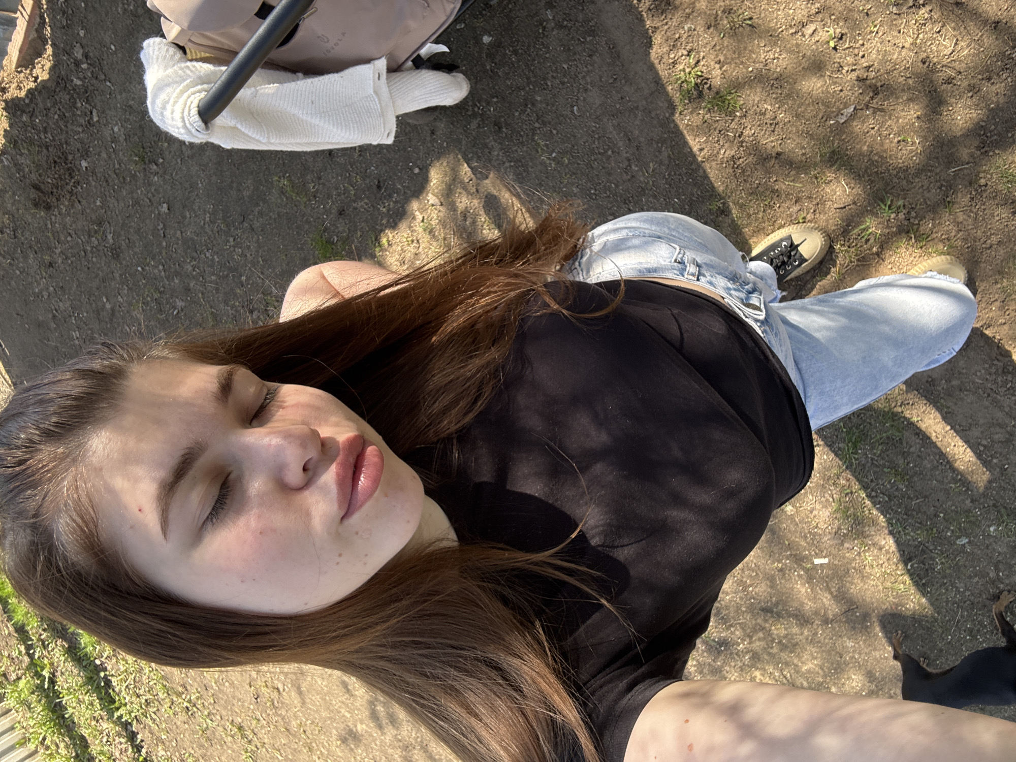 A woman lies on the ground, angled towards the camera, with her eyes closed and lips visible. She has long brown hair. She is wearing a black shirt and light blue jeans. Part of a baby stroller is in the background, along with a white knitted item. Her shoes are visible. A black dog is partially in the frame, near her feet. The scene appears to be outdoors, possibly in a park or grassy area, with dirt and scattered grass. There is sunlight. There is no definitive evidence in the image to identify the location as Мещёры, Russia.