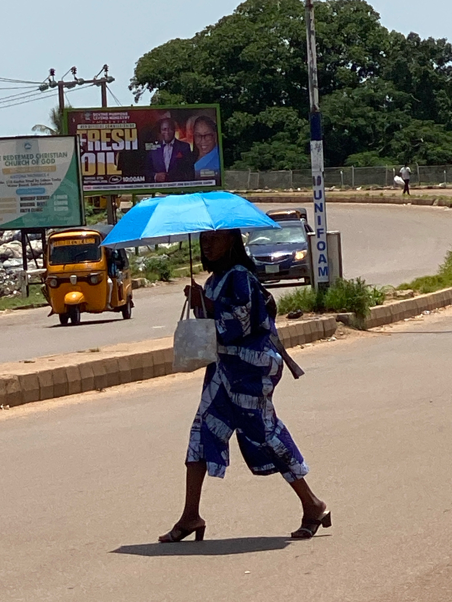 A woman walks across a paved road in Sabon Tasha, Nigeria, holding a blue umbrella over her head. She is wearing a blue and white patterned outfit and dark open-toed heeled sandals. She carries a white tote bag in her left hand and a dark object, possibly a rolled-up mat or umbrella, tucked under her right arm. The street is lined with a concrete curb and has a sandy shoulder. In the background, a yellow autorickshaw is visible on the road to the left, and a dark blue car is approaching from behind the woman. A utility pole with vertical lettering stands on the right side of the road. Further in the background, billboards, trees, and distant figures can be seen. The sky is bright and appears to be daytime.