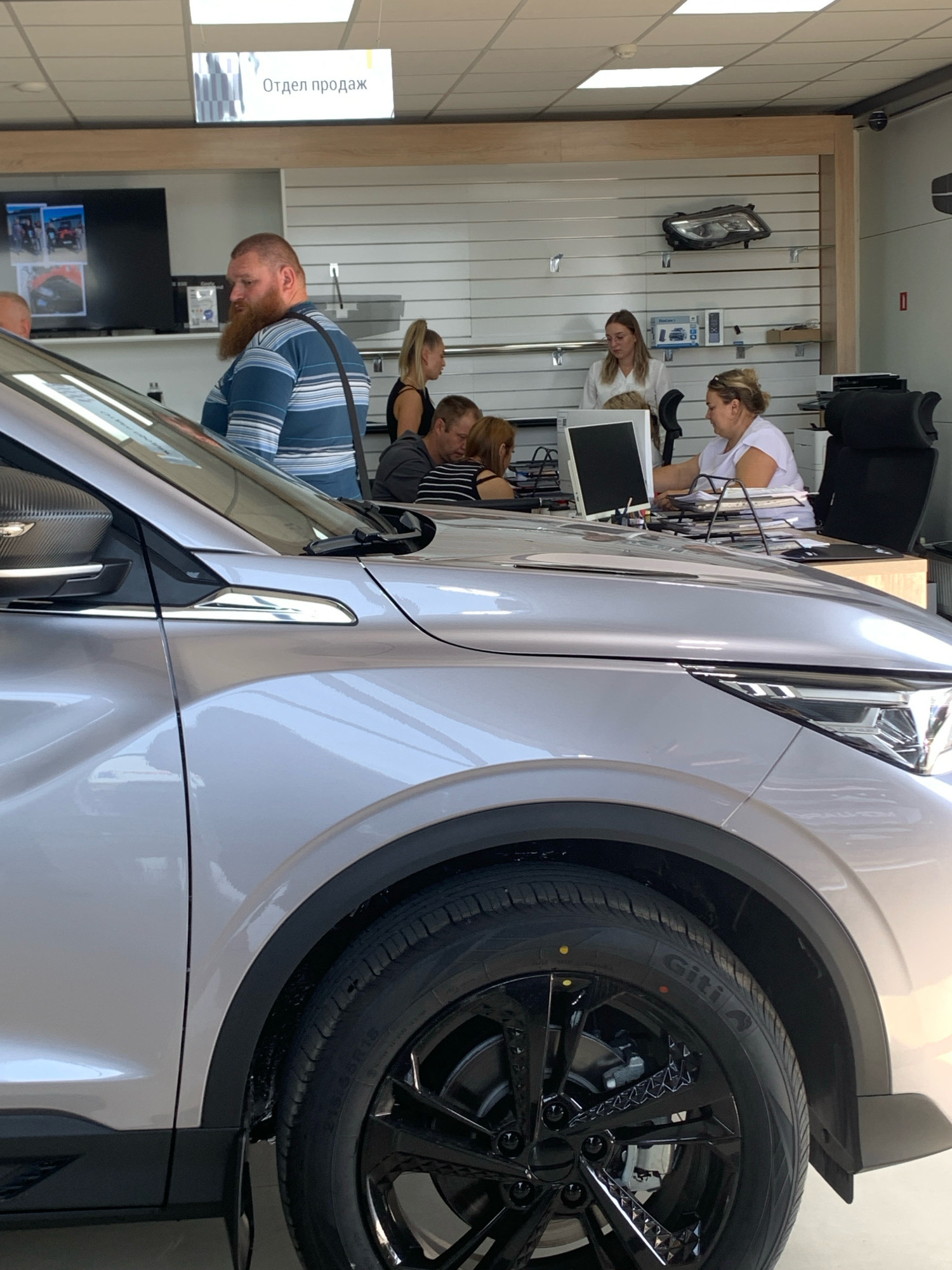 In a brightly lit car dealership showroom, a silver SUV is prominently displayed in the foreground. Several people are visible in the background, interacting in what appears to be a sales or administrative area. A man with a beard and a blue and white striped shirt stands near the car. To his right, a woman with blonde hair pulled back in a ponytail is seen. Further to the right, two women are seated at desks with computers and papers. One woman, wearing a white top, is engaged with a computer, while another, with blonde hair tied back and wearing glasses, looks towards a monitor. On the wall behind them, a car headlight is displayed on a shelf. A sign overhead reads "Отдел продаж," which translates to "Sales Department." The overall environment suggests a typical car dealership setting, possibly in the vicinity of Чыжэвіцкі сельскі Савет, Belarus, given the signage in Russian.