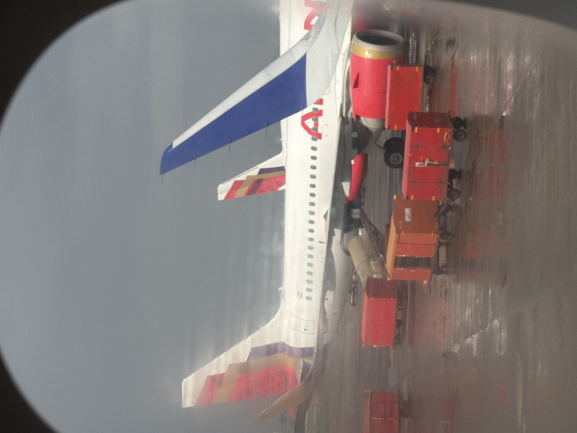 The image captures a scene at an airport, likely in Mumbai, India, given the presence of an IndiGo aircraft. Two airplanes are visible. The primary aircraft, an IndiGo Airbus A320 family aircraft, is white with red accents on its engines and tail. The tail features a colorful geometric design in shades of red and purple. The second aircraft, partially obscured, has a blue and white tail fin. Several baggage carts are positioned on the tarmac, some are red, one is orange, and one is a lighter tan color. The tarmac appears wet, suggesting recent rain or a damp environment. The sky is overcast and gray, contributing to a subdued atmosphere. The image is framed by the dark oval shape of an airplane window. No people are clearly visible in the frame, suggesting the ground crew may be out of shot or the aircraft is parked and unattended at this moment. The scene conveys a sense of operational readiness at the airport, with baggage handling equipment in place for servicing the aircraft.