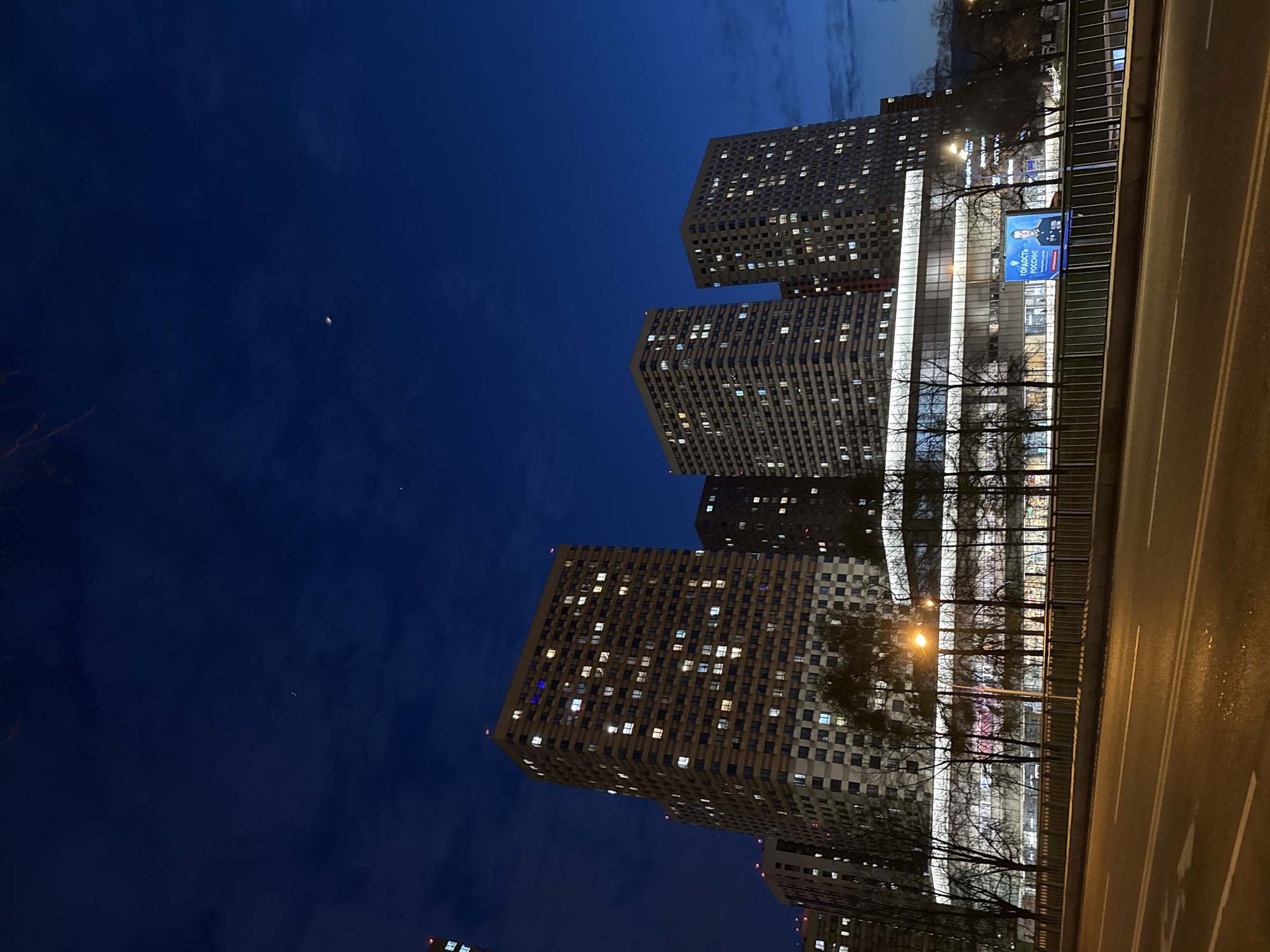 This image captures a nocturnal urban landscape in Khimki, Russia, featuring tall residential buildings, a road, and a dark blue sky. Several prominent high-rise buildings, likely apartment complexes, rise vertically, displaying a grid of numerous windows, many of which are illuminated with warm light, suggesting active occupancy. The architectural style appears modern and functional.
A multi-lane road or bridge occupies the right side of the frame, stretching diagonally with visible white and yellow lane markings. The road surface appears wet and reflective, possibly from recent rain, mirroring the city lights. A metal barrier or fence separates the road from the area closer to the buildings. Between the road and the towering structures, bare trees stand with their intricate branches silhouetted against the brighter background of building facades and ambient light. A streetlight casts a warm, orange glow near the road.
In the mid-ground, a large blue electronic billboard or display is visible. It features the portrait of a man, likely a public figure, and prominently displays the Russian text "ГОРДОСТЬ РОССИИ!", which translates to "PRIDE OF RUSSIA!".
The sky, dominating the upper left, is a deep, dark blue with faint cloud patterns. A small, bright crescent shape, possibly a sliver of the moon or another celestial object, is visible in the upper left quadrant. The scene depicts a typical late evening or nighttime view of a bustling city, characterized by artificial lights under a calm, dark sky. No specific human activity is the primary focus; rather, it is a static urban snapshot.