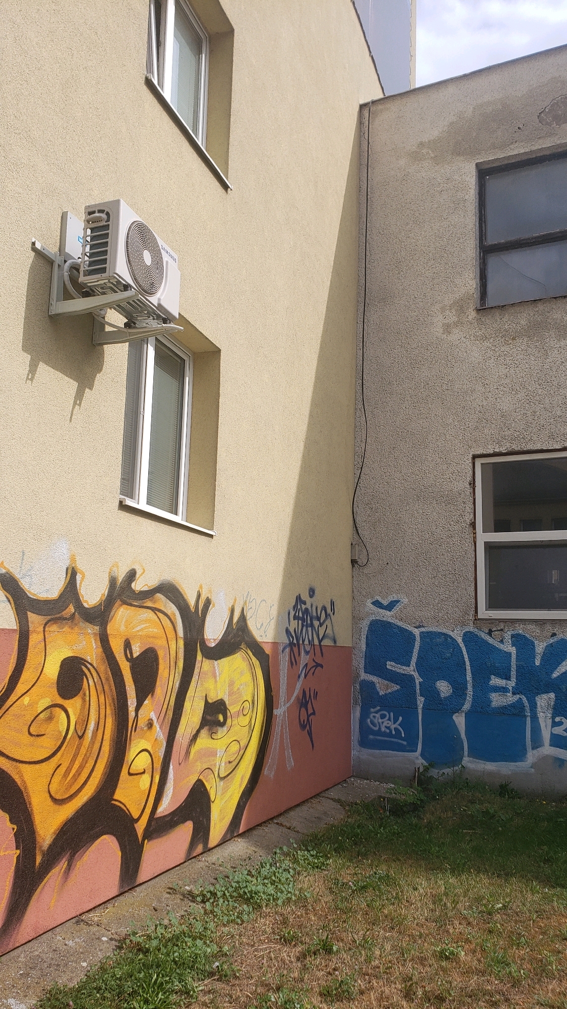 The image depicts the exterior of two adjacent buildings, one with a light yellow facade and the other with a beige, textured facade, situated in the city of Prešov, Slovakia. The yellow building features an air conditioning unit mounted on the wall below a window. Both buildings have windows; the yellow building has a tall, narrow window with blinds, while the beige building has two rectangular windows with dark panes. The lower portions of both buildings' walls are covered in graffiti. On the left, there is graffiti in yellow and black with orange accents, featuring abstract, bold lettering. To the right of this, on the beige wall, blue graffiti with white accents is visible, also with blocky lettering. A narrow strip of dry grass and some green weeds is present at the base of the buildings. A shadow from an unseen object falls diagonally across the yellow building.