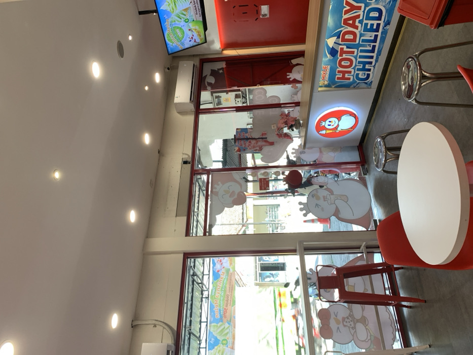 The image shows the interior of a store in Kecamatan Piyungan, Indonesia, located at coordinates -7.837, 110.474. The store is brightly lit with a white ceiling containing several recessed lights. The walls are painted white and red, and the floor is tiled. There are large glass windows and doors that open to an exterior view of a residential area. The store features several cartoon images of snowmen and a woman, all wearing crowns. The front counter is decorated with a large sign that reads "Hot Day Chilled." There are three white plastic chairs and two silver metal stools situated around a white round table. A red barstool is next to the window. A black and white television is hanging on the red wall, showcasing a promotional image of a dessert. The store is empty and unoccupied.