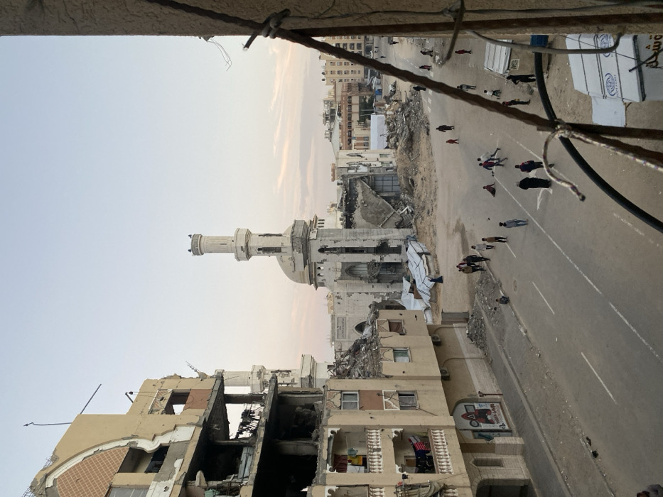 A war-torn street features heavily damaged buildings and a mosque with a visible minaret. Rubble and debris litter the area. People are walking along the road, seemingly navigating through the devastation. Buildings in the background appear intact. The view is taken from an elevated position, perhaps a building's upper floor, with some foreground elements suggesting a balcony or ledge.