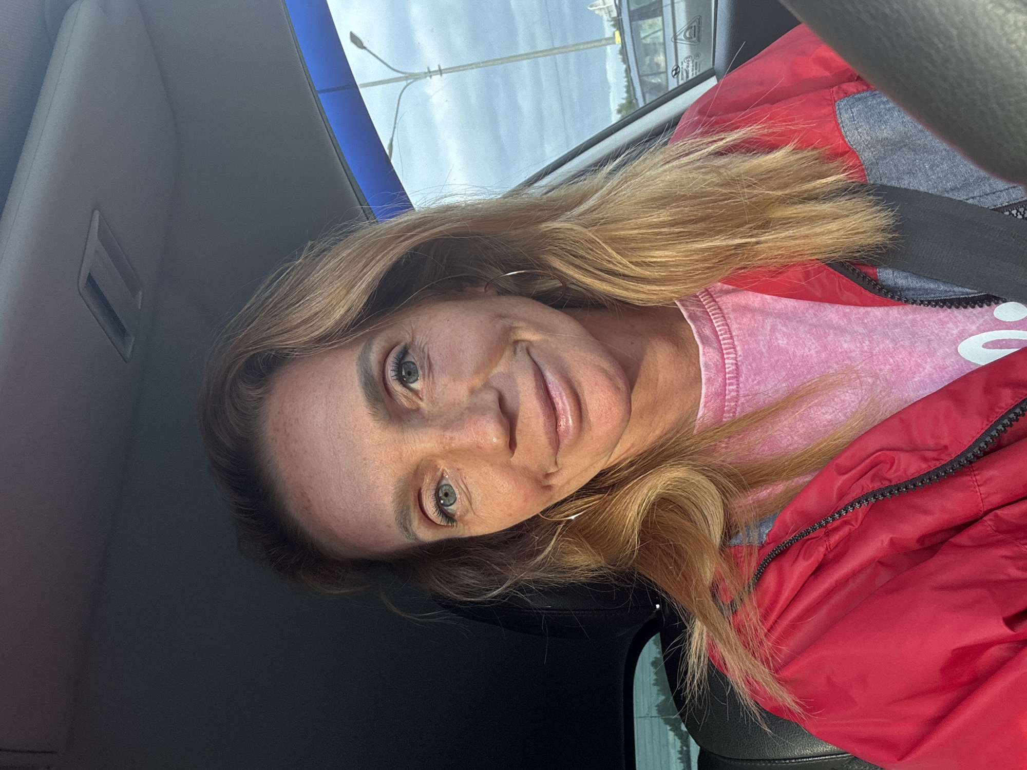 A woman is captured in a selfie taken from inside a car. She is wearing a red jacket over a pink t-shirt and has blonde hair. Her eyes are blue, and she is looking at the camera with a slight smile. The background shows the interior of a car, with a glimpse of the sky and a street light visible through the window. The photograph was taken in Kolyasovo, Russia.