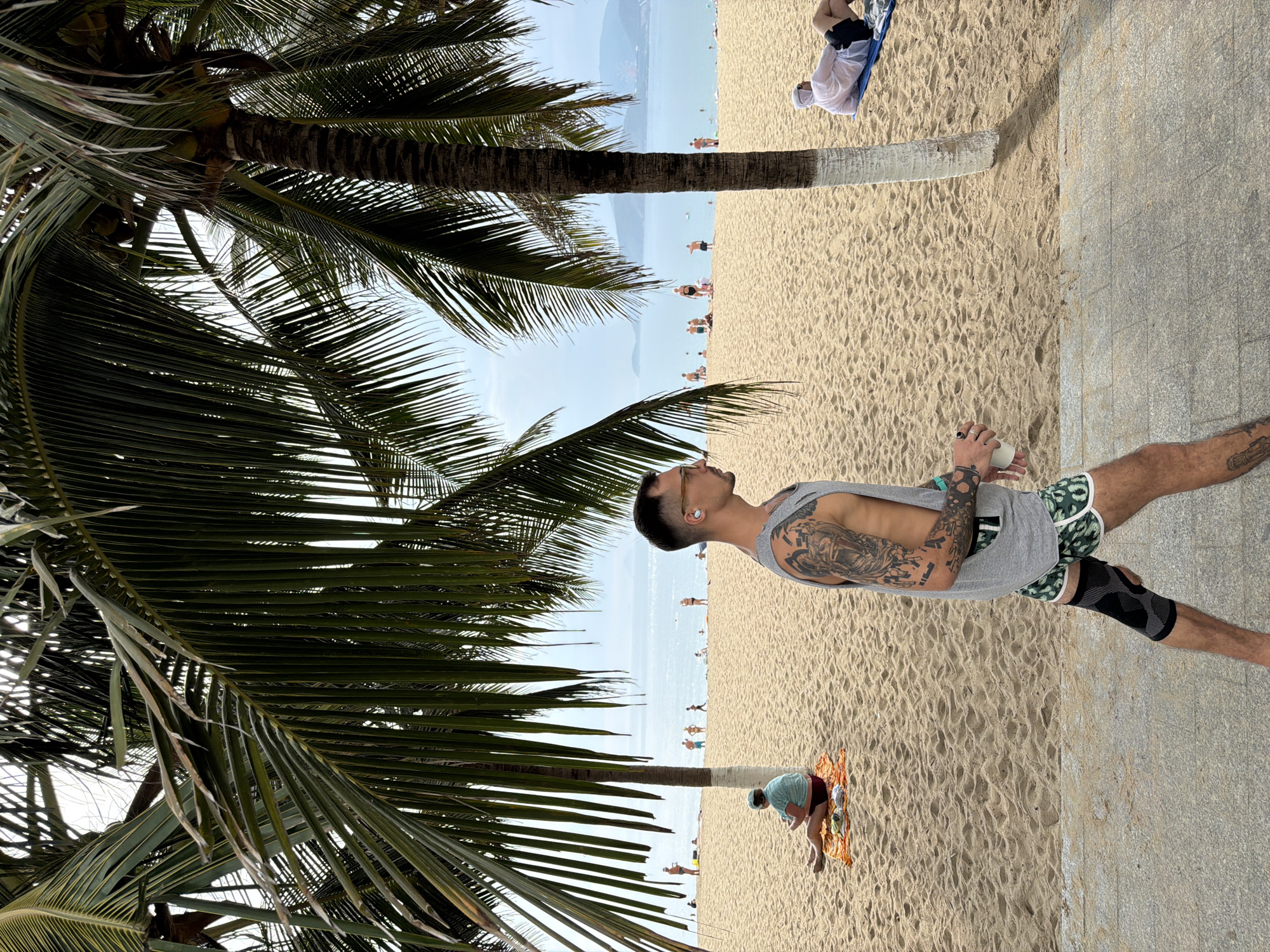 A man stands on a paved walkway adjacent to a sandy beach in Nha Trang Ward, Vietnam. The man, wearing a gray tank top, patterned shorts, and a knee brace on one leg, holds a white cup. He has tattoos on his arms and an earpiece. Palm trees frame the scene, with some leaves obscuring the sky. The beach has other people on it, some lying down and others walking. The sea is in the background under a hazy sky.