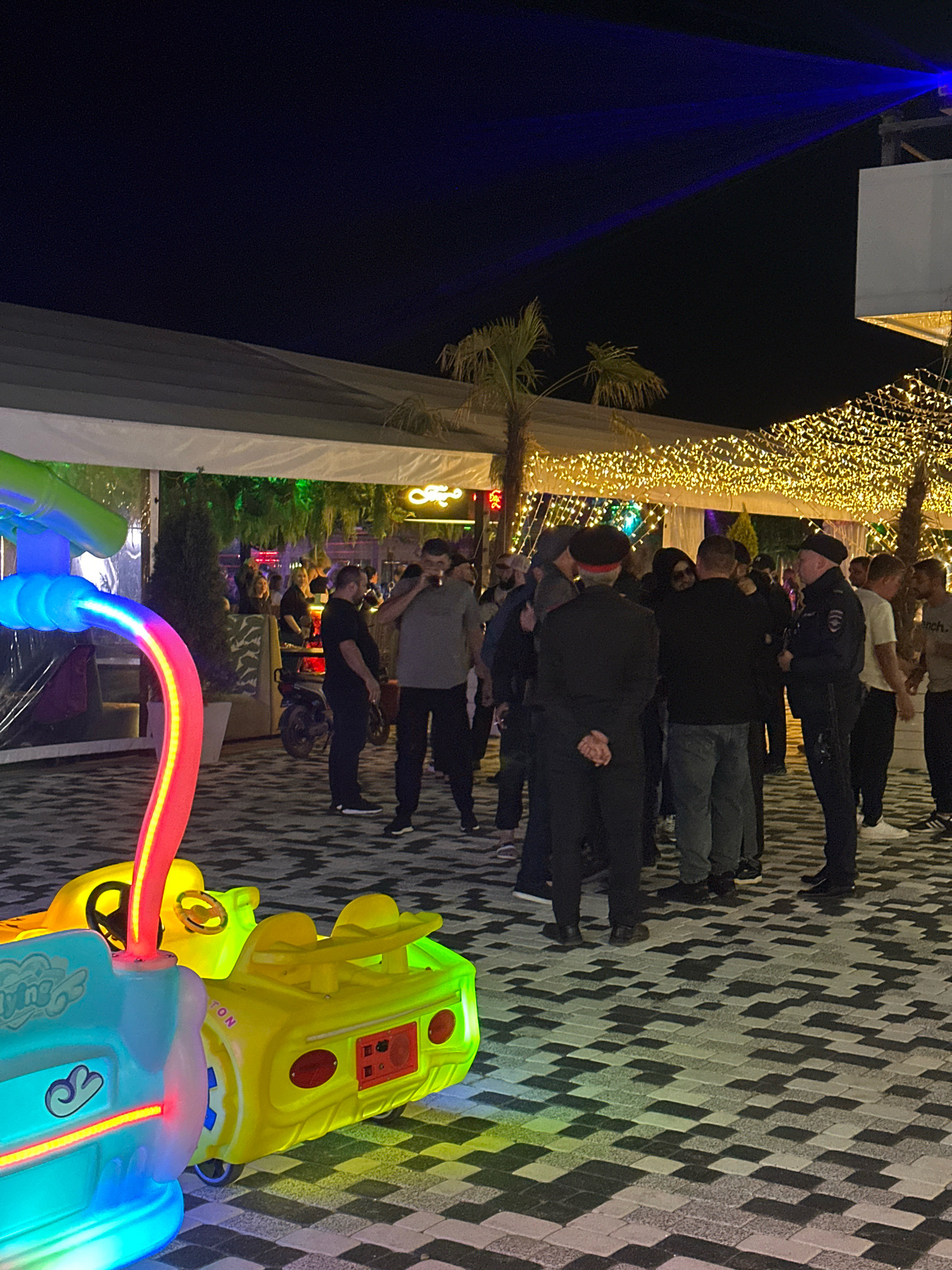 An outdoor, paved area at night in Lermontovo, Russia, is illuminated by string lights overhead and neon lights on a colorful children's ride in the foreground. Several people are gathered, some appearing to be in conversation. Two individuals in uniform, one in a police uniform, are also present. A parked motorcycle is visible in the background. The scene suggests a public gathering or an event taking place at an entertainment venue.