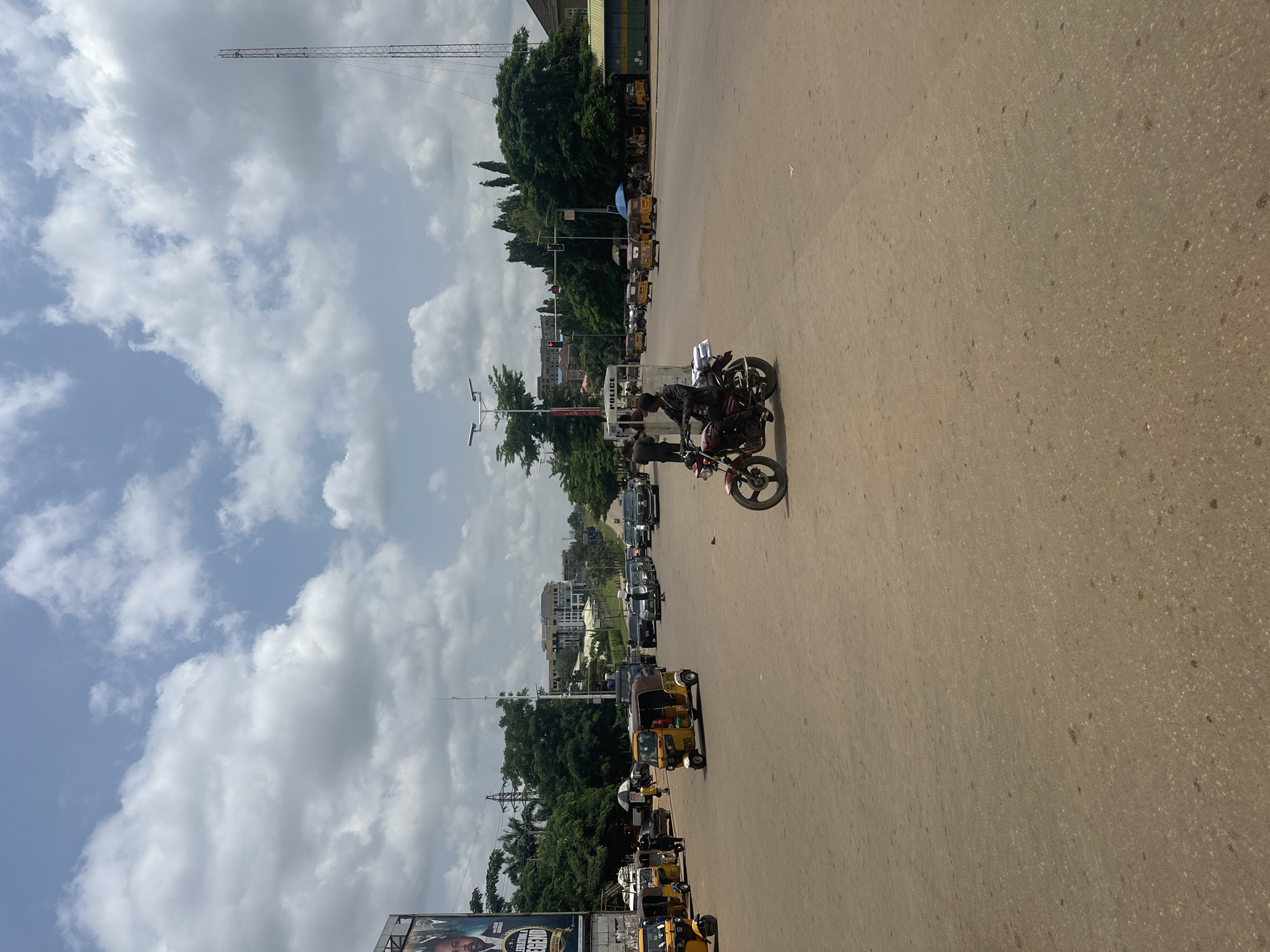 The image depicts a street scene in Abuja, Nigeria, likely a busy intersection with moderate traffic. In the foreground, a man is riding a red motorcycle. Behind him, several yellow auto-rickshaws, commonly known as Keke Napep, are parked or moving. Further in the background, various cars and other vehicles are visible, suggesting ongoing movement. A "POLICE" booth is situated on the left side of the road, with a police officer standing near it. The environment includes trees, buildings, and a tall communication tower in the distance. The sky is partly cloudy. A large billboard featuring a man's face with the text "CELEBRATING LEGEND" is present on the left edge of the image. The road surface appears to be paved and sandy.