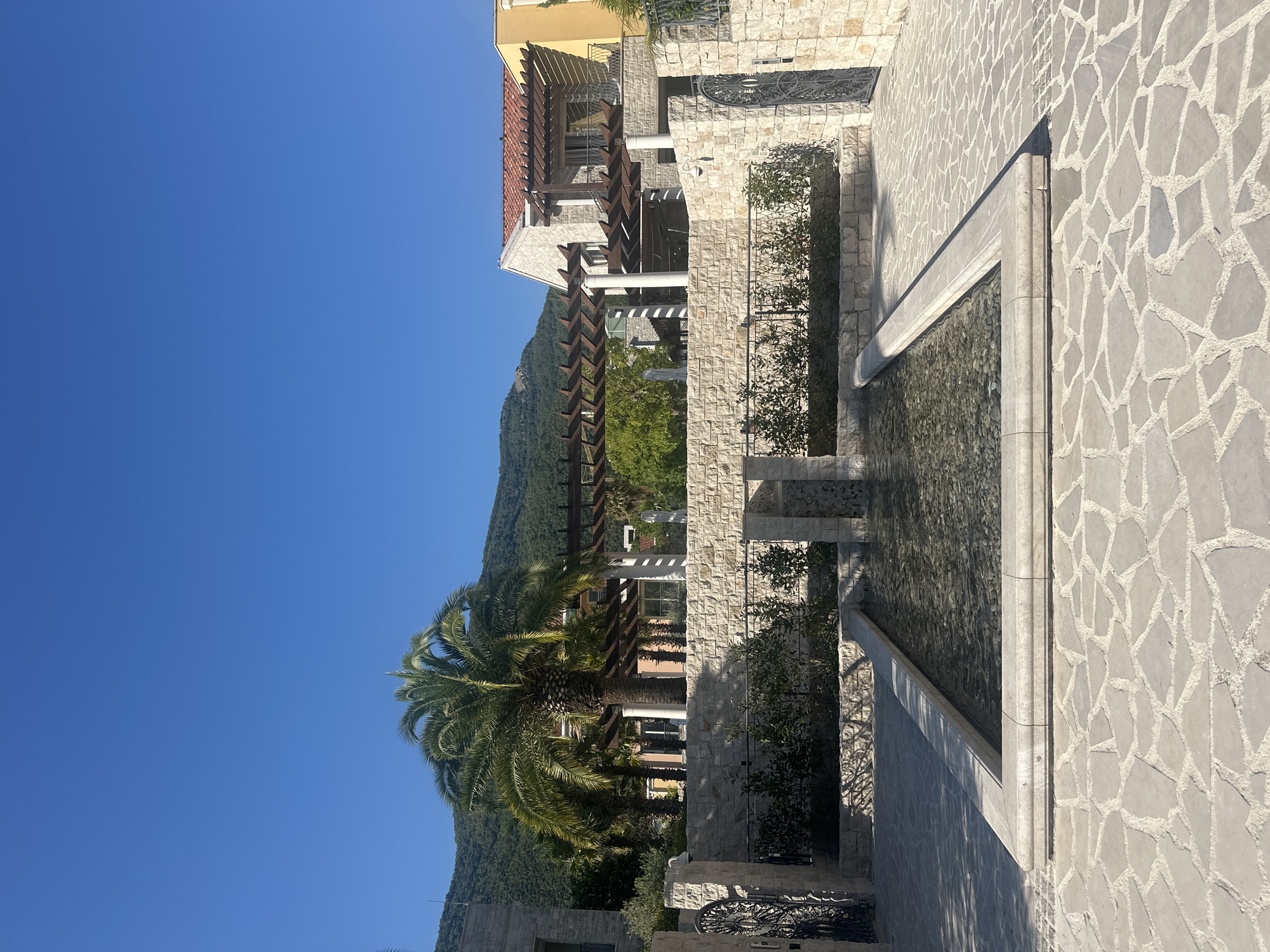 The image depicts a sunny outdoor scene in Kumbor, Montenegro, featuring a modern courtyard with a rectangular water feature at its center. The water feature has a stone border and is filled with clear water containing submerged pebbles. The courtyard is paved with irregular, light-colored flagstones.
Behind the water feature, a low stone wall topped with a dark metal fence encloses a strip of greenery. Beyond the wall, a large palm tree stands prominently, with its fronds casting shadows. In the background, there are contemporary buildings with light-colored stone facades and red tile roofs, along with wooden pergolas. A lush, tree-covered hillside rises behind the structures, under a clear blue sky.
To the left and right of the central area, there are ornate black metal gates set within stone pillars, suggesting an entrance or boundary. The overall impression is of a well-maintained, upscale residential or resort-like setting in a Mediterranean climate. There are no people visible in the image.