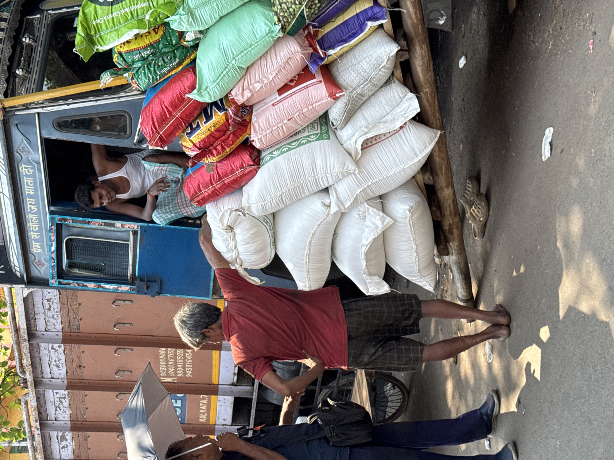 The image captures a scene in Kolkata, India, where a delivery truck is heavily loaded with sacks of goods, primarily appearing to be food items. One man is sitting in the driver’s seat. Another man is standing beside the truck, seeming to interact with the sacks. A third person is walking near the truck, holding an umbrella. The truck has the word "KOLKATA.7" and several phone numbers printed on it, and its sign has Hindi text. The sacks, stacked high, are of various colors and brands. The scene is on a street, with the sunlight casting shadows.