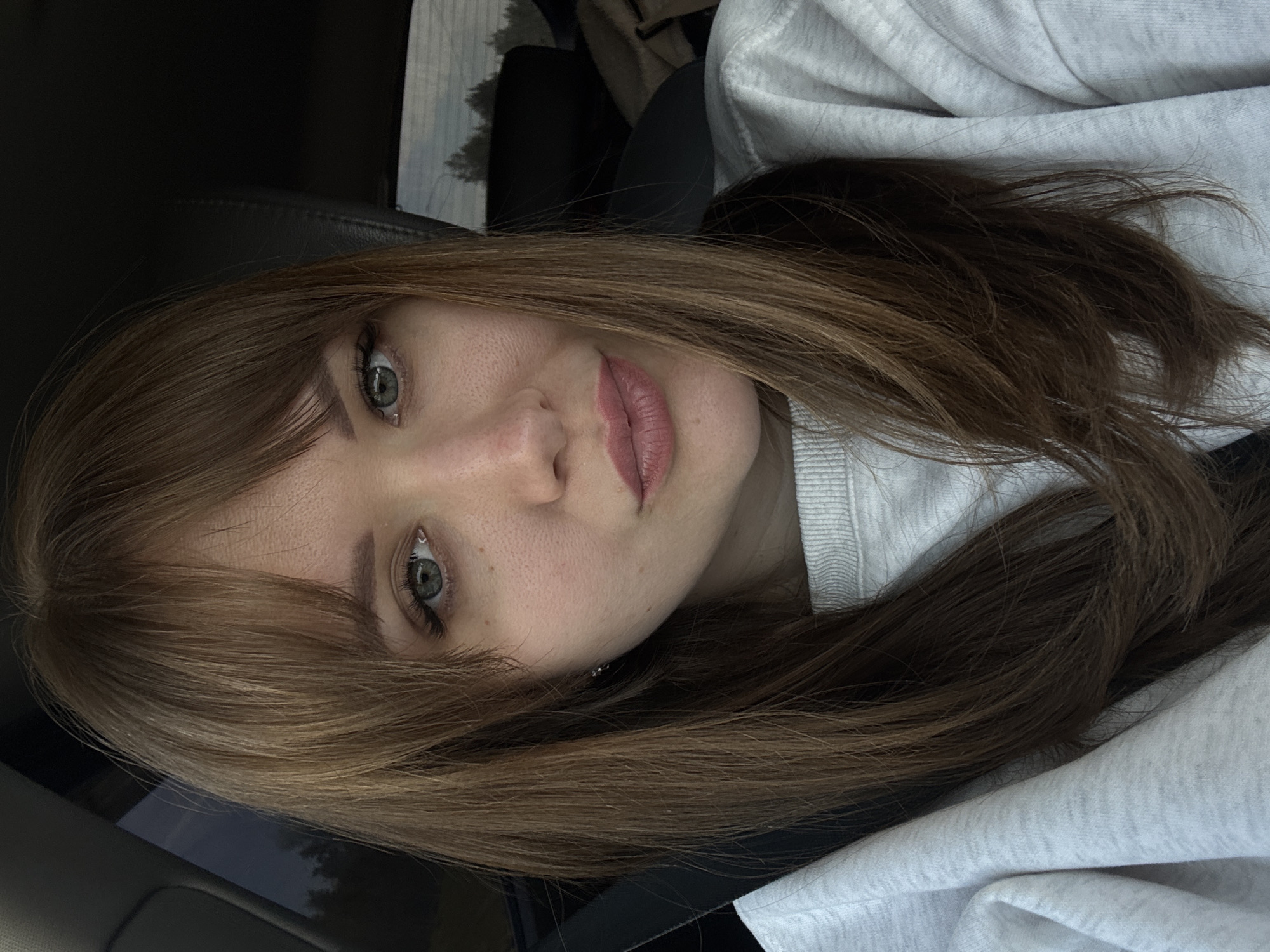 A selfie of a young woman with light brown hair and green eyes, likely taken in a car. She's wearing a light gray sweatshirt. The background shows the car interior and a glimpse of the outside through the window. No evidence suggests it's specifically in the city of Yegoryevo, Russia.