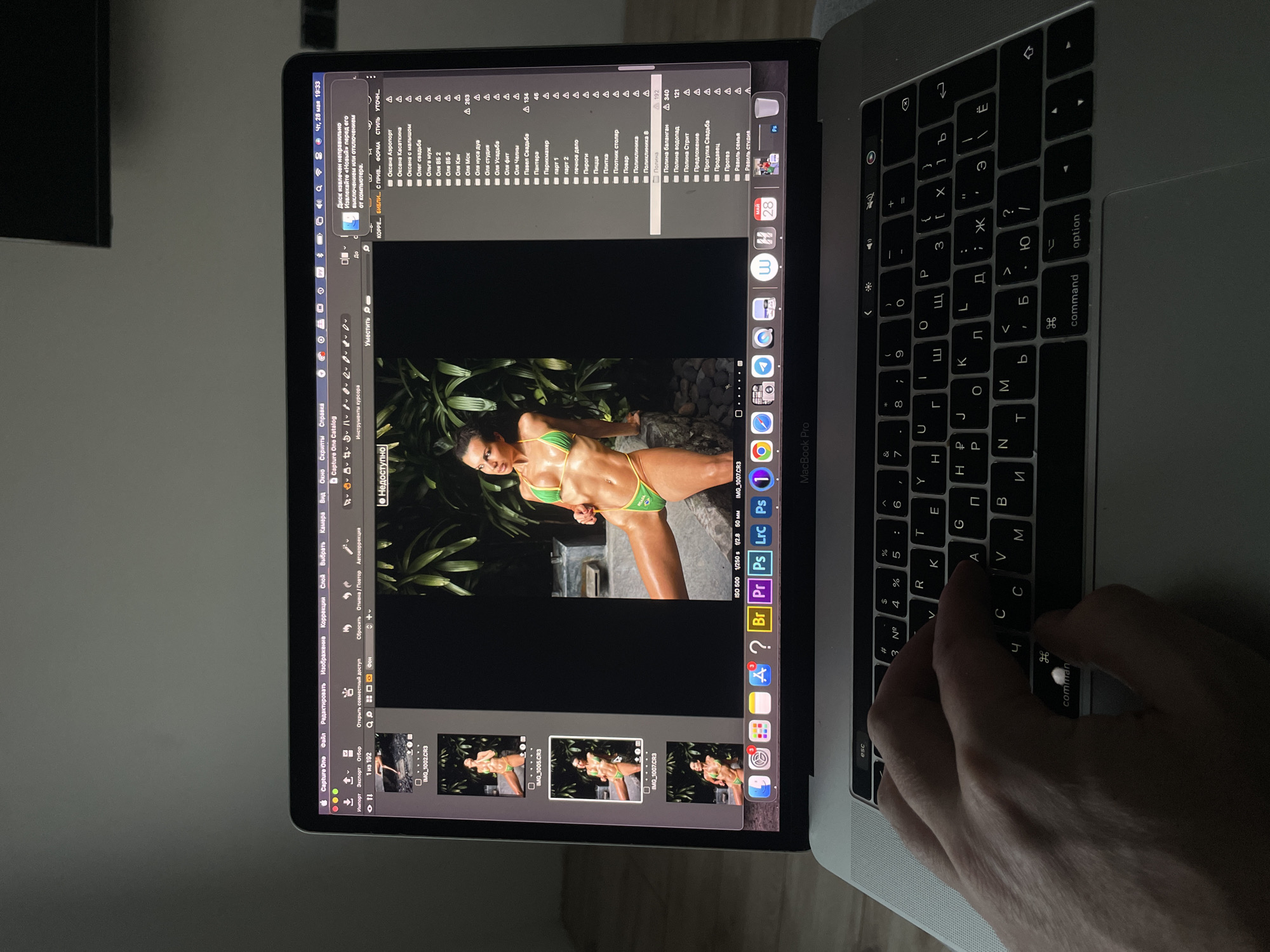 The image shows a person working on a MacBook Pro laptop. The laptop is open, displaying a photo editing program, Capture One. The editing program shows a photo of a woman in a green bikini, surrounded by tropical foliage. The laptop keyboard is Cyrillic, suggesting the location is likely Russia, or another country that uses Cyrillic script. A hand is visible on the right side of the keyboard, indicating the person is typing or navigating. The folders in the Capture One catalog feature Russian names, for example Оксана Касаткина, which confirms that it is a Russian language catalog, and may confirm that the location is Russia.
There is no evidence in the image to suggest it is in the city of Kazan, Russia.