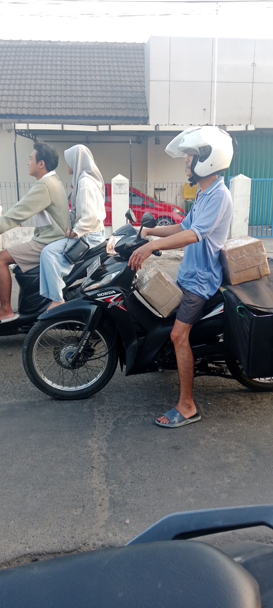 The image shows three individuals on two motorcycles on a street in Maguwoharjo, Indonesia. The foreground is dominated by the side view of a black Honda motorcycle with a delivery bag and two packages strapped to it. The rider, a male wearing a blue polo shirt, grey shorts, and a white helmet, is in profile, looking forward. Behind him, on the same motorcycle, is a female wearing a light grey hijab and a white knitted sweater, also looking forward. To the left, on another motorcycle that is partially visible, a male wearing a light-colored jacket and beige shorts is looking to the left. The background includes a building with a tiled roof and a modern facade, a red car, and a blue metal fence. The road surface is asphalt.