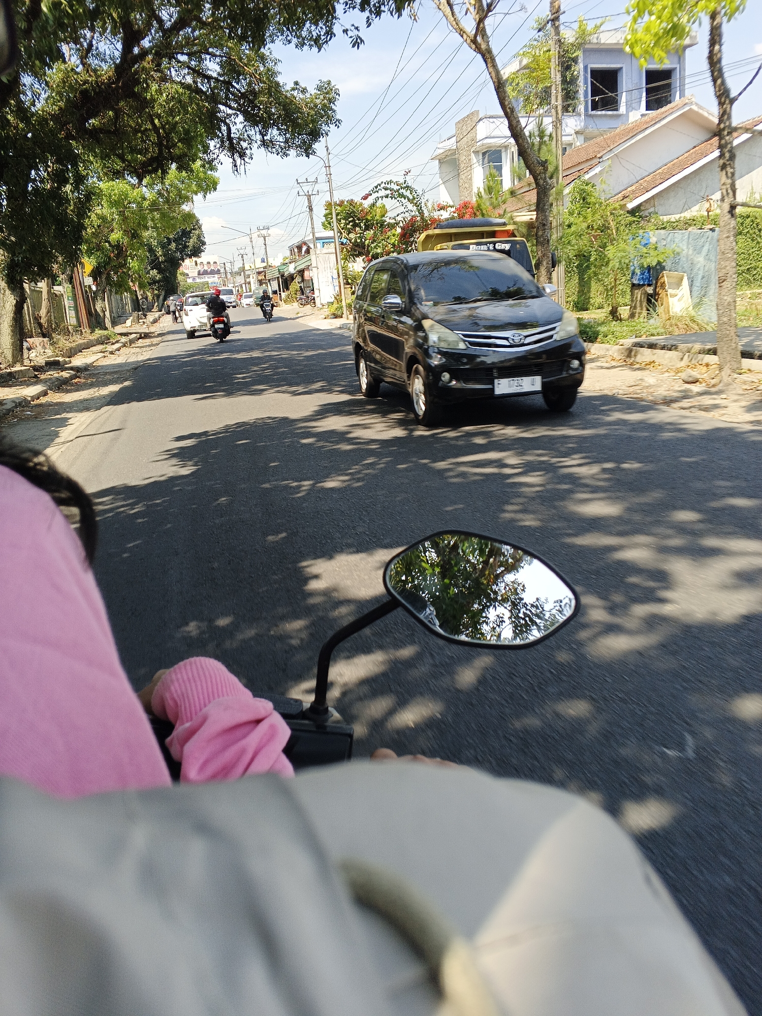 The image shows a view from the perspective of someone riding on the back of a motorcycle in Sukabumi, Indonesia. The rider's pink sleeve is visible in the foreground. In front of the motorcycle, a black car is driving on an asphalt road lined with trees and houses. The car has a license plate that appears to read "F 9732 U." The road is partially shaded by the trees, creating dappled light patterns on the asphalt. The sky is clear and blue. In the distance, there are several buildings, including a house with a light blue upper story and an orange tiled roof. The overall environment suggests a sunny day in a suburban or semi-urban area.