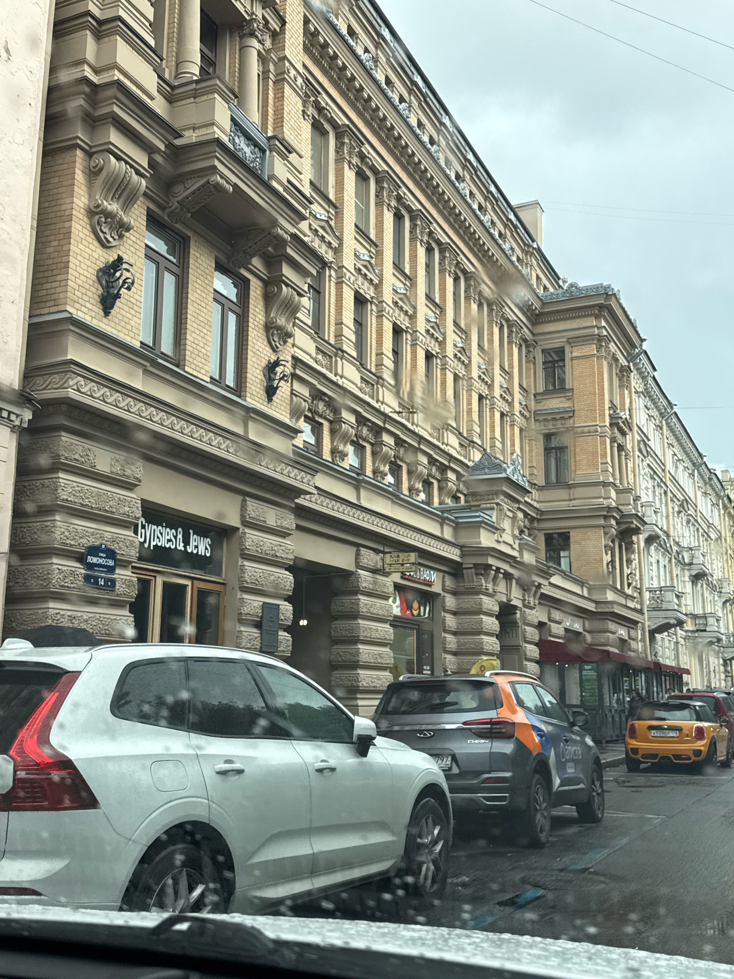 The image depicts a street scene in Saint Petersburg, Russia, on a cloudy day. The focus is on the ornate architecture of buildings lining the street, characterized by their yellow brick facades, numerous windows, decorative cornices, and classical architectural elements such as columns and elaborate carvings. In the foreground, several cars are parked and driving. A white Volvo SUV is prominently visible on the left, partially obscuring the building. Further down the street, a gray and orange Chery SUV and a yellow Mini Cooper are also present. The storefronts of the buildings are visible, with one sign reading "Gypsies & Jews" above a doorway. Another sign, in Cyrillic, appears to be "Ломоносова" (Lomonosova), indicating the street name. The wet pavement suggests recent rain. The overall impression is of a historic urban environment, likely a commercial or residential district in Saint Petersburg.