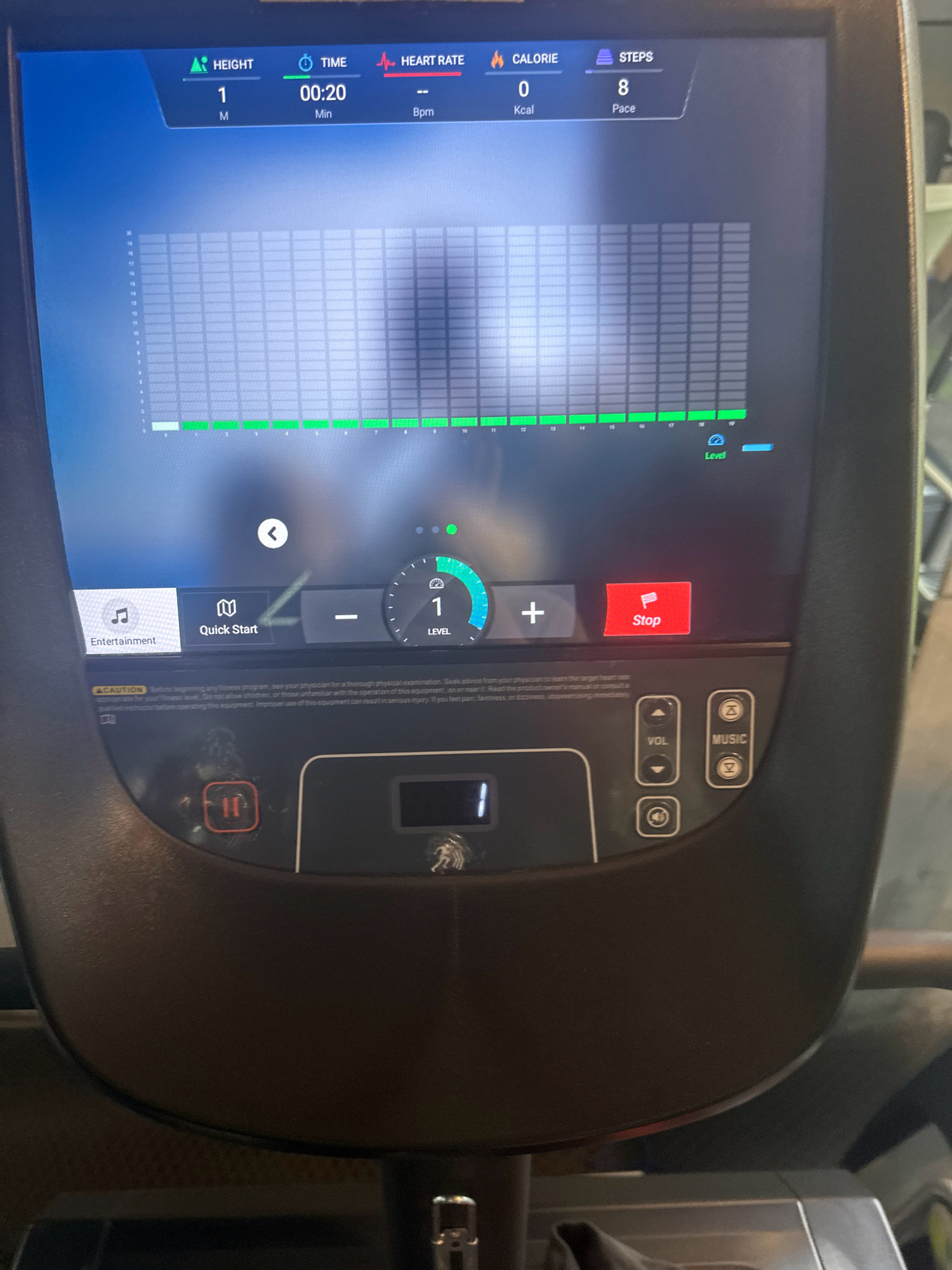The image shows a close-up view of a modern fitness machine's console, likely a treadmill or elliptical, in what appears to be an indoor gym setting. The console displays various workout metrics on a digital screen, including "HEIGHT" at "1 M", "TIME" at "00:20 Min", "HEART RATE" at "0 Bpm", "CALORIE" at "0 Kcal", and "STEPS" at "8 Pace". A bar graph runs across the screen, showing a green progression from left to right, indicating activity. Below the graph, there are control buttons for "Entertainment", "Quick Start", "Level" adjustment (indicated by a dial showing "1"), and a "Stop" button. There are also buttons for volume control ("VOL") and music selection ("MUSIC"), as well as a "Pause" button with a red outline and "II" symbol. The machine console itself is black and appears to be integrated into a larger piece of fitness equipment. In the background, parts of another exercise machine and possibly a person are visible, but they are out of focus. There is no indication of the specific location being Lalitpur, Nepal.