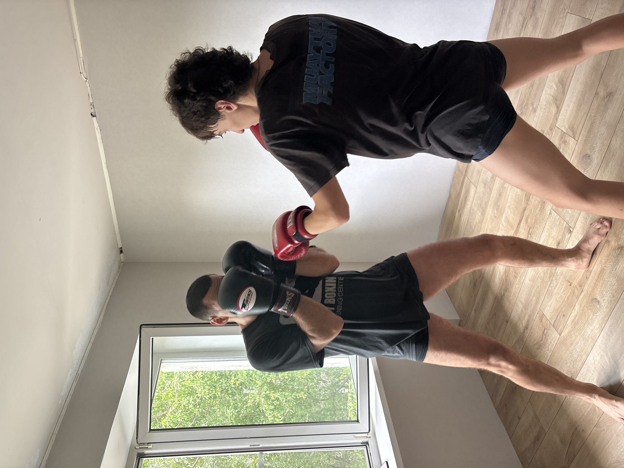 The image displays two individuals, likely men, engaged in a combat sports training session within an indoor facility. The scene is illuminated by natural light entering through a large window on the left, revealing green foliage outside, indicating a daytime setting, possibly in spring or summer. The room features light-colored walls and a light wooden floor, suggesting a functional training environment.
In the foreground, one participant with short dark hair and a beard is shown in a defensive stance, hands raised to protect his head. He wears a dark t-shirt with the partial text "BOXIN TRAINING CENTE" and black shorts. He is equipped with black boxing gloves bearing the "TWINS" brand logo and red details. Both individuals are barefoot.
Behind him, the second participant, who has curly dark hair, is in an active or offensive posture, wearing red boxing gloves. His dark t-shirt clearly displays "MUAY THAI FACTORY" across the back, and he also wears black shorts. Their postures suggest a focused practice or sparring session.
The setting, possibly in Chaikovsky, Russia, appears minimalist, with no additional furniture or decorative elements visible, emphasizing its purpose as a training space. Some electrical conduits are visible near the ceiling.