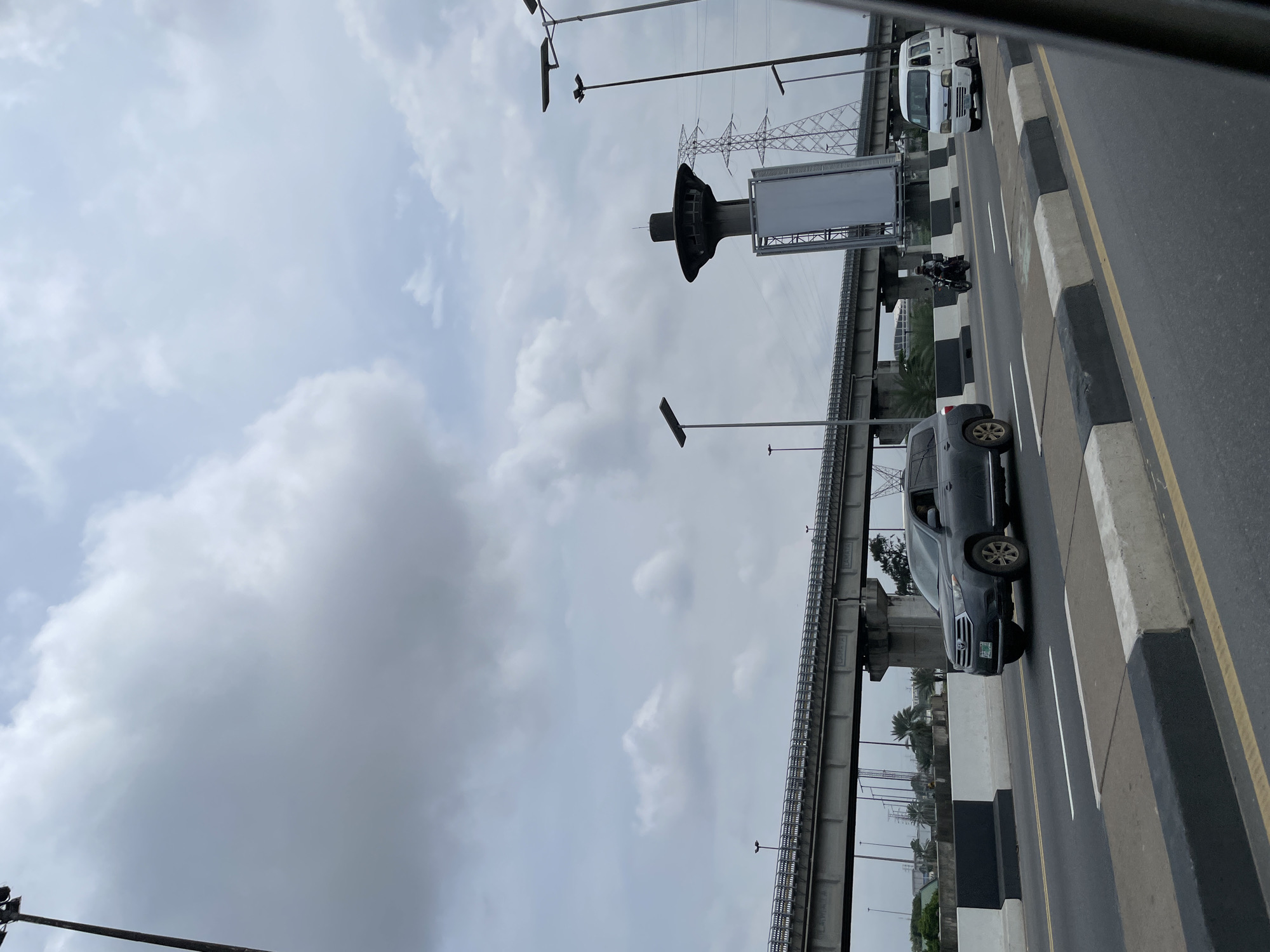 This is a mid-distance, eye-level shot taken from a moving vehicle on a highway in Lagos, Nigeria. The sky is heavily overcast with dramatic grey clouds, suggesting a humid or rainy day. In the foreground, the road is paved with asphalt and divided by a yellow line. The sides of the road are bordered by concrete barriers painted with black and white stripes.
Dominating the middle ground is a multi-lane highway or flyover system, supported by concrete pillars. The elevated structure stretches across the frame. On the highway, several vehicles are visible: a dark grey SUV is in the left lane, and a white minivan is in the right lane. A person on a motorcycle is also seen traveling between the lanes.
In the background, a distinctive tall, circular structure with a flared top, resembling a control tower or observation deck, stands prominently. Adjacent to it are tall electricity pylons, characteristic of urban infrastructure. Sparse tropical vegetation, including palm trees, can be seen peeking out from beneath the elevated highway. The overall impression is one of a busy, modern urban environment under a somber sky.