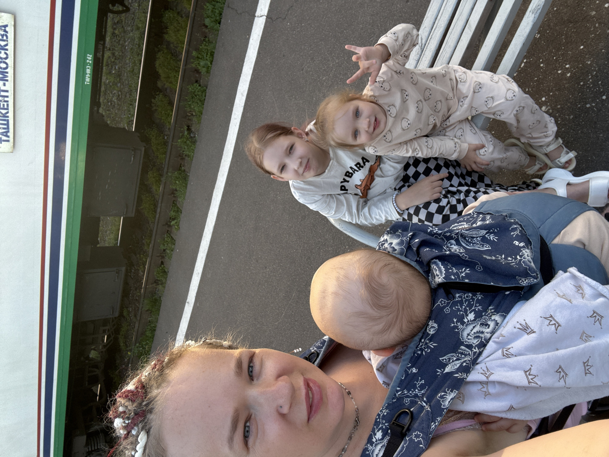 A woman is taking a selfie at a train station in Uzunovo, Russia. She is wearing a patterned baby carrier with a baby inside. To her right are two girls sitting on a bench. The older girl is wearing a white shirt with "PYBARA" printed on it, and checkered pants. The younger girl is wearing a pajama set with teddy bear prints. The train is visible in the background with "ТАШКЕНТ-МОСКВА" written on it.