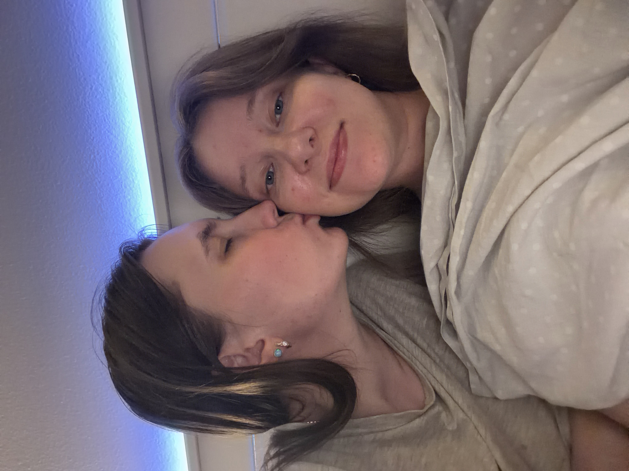 Two individuals, presumably a couple, are depicted in a close-up, horizontal shot. One person is leaning in to kiss the other on the cheek. They are both under a light-colored duvet with a subtle white polka dot pattern. The individual being kissed is looking towards the camera with their eyes open and a slight smile. The person kissing has their eyes closed. Both individuals have light skin and brown hair.
The setting appears to be indoors, possibly a bedroom, suggested by the presence of a duvet and soft lighting. A bright blue light source is visible on the left side of the frame, casting a cool glow. The overall mood is intimate and affectionate. The image is likely taken during the evening or night, given the subdued lighting and the cozy atmosphere. No text is visible in the image. The location context is Portland, United States of America.