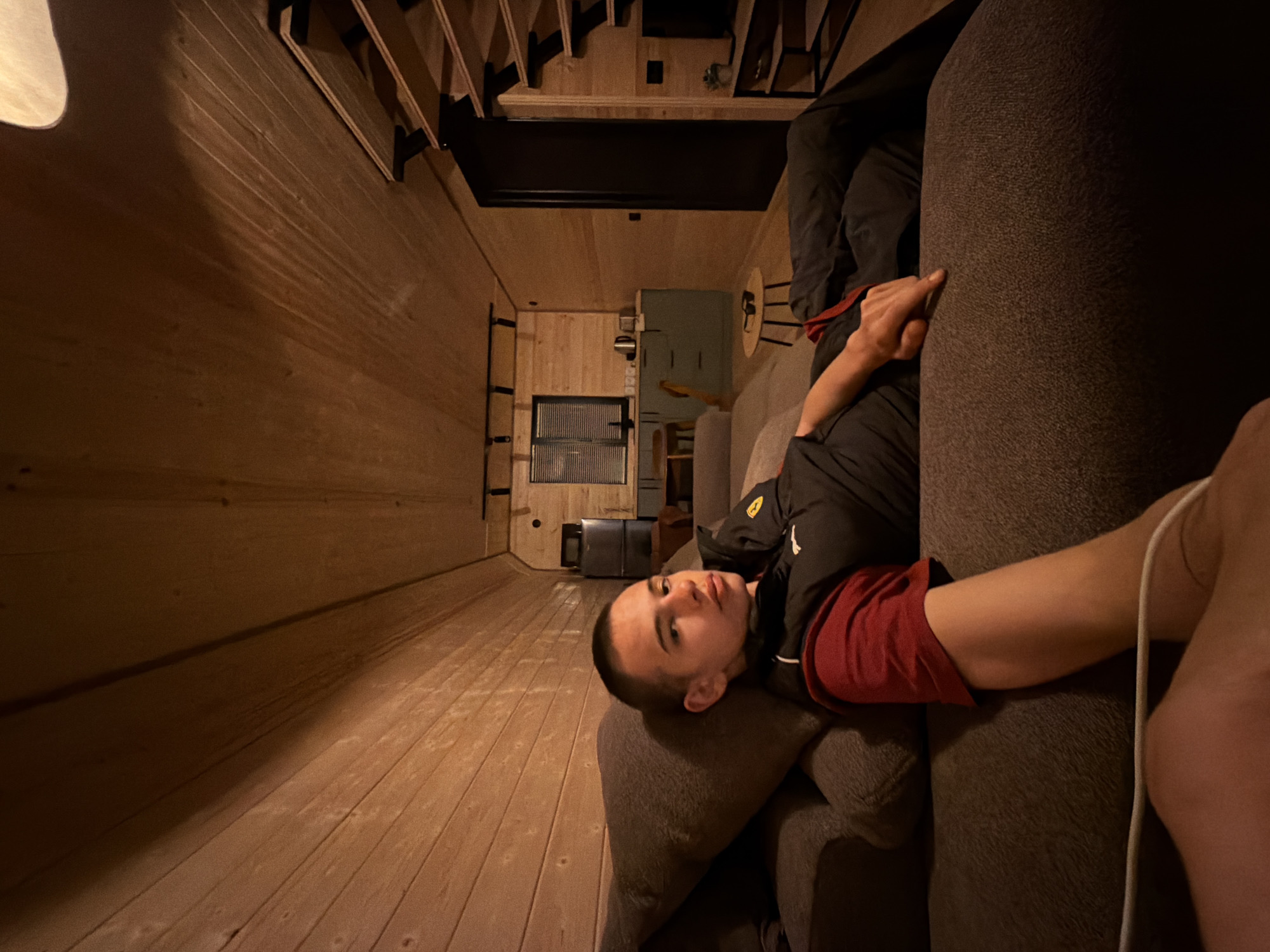 A young male with a buzz cut is lying on a sofa in a room with wooden paneled walls and ceiling. He is wearing a black and red sports top. His left arm rests on the sofa arm, and his right arm is extended across his torso. In the background, a small kitchen area with light blue cabinets and a window with blinds is visible. A set of stairs leading to an upper level is on the right side of the room. The room is dimly lit, with warm light illuminating the wooden surfaces. A small round table with sunglasses on it is positioned in front of the sofa.