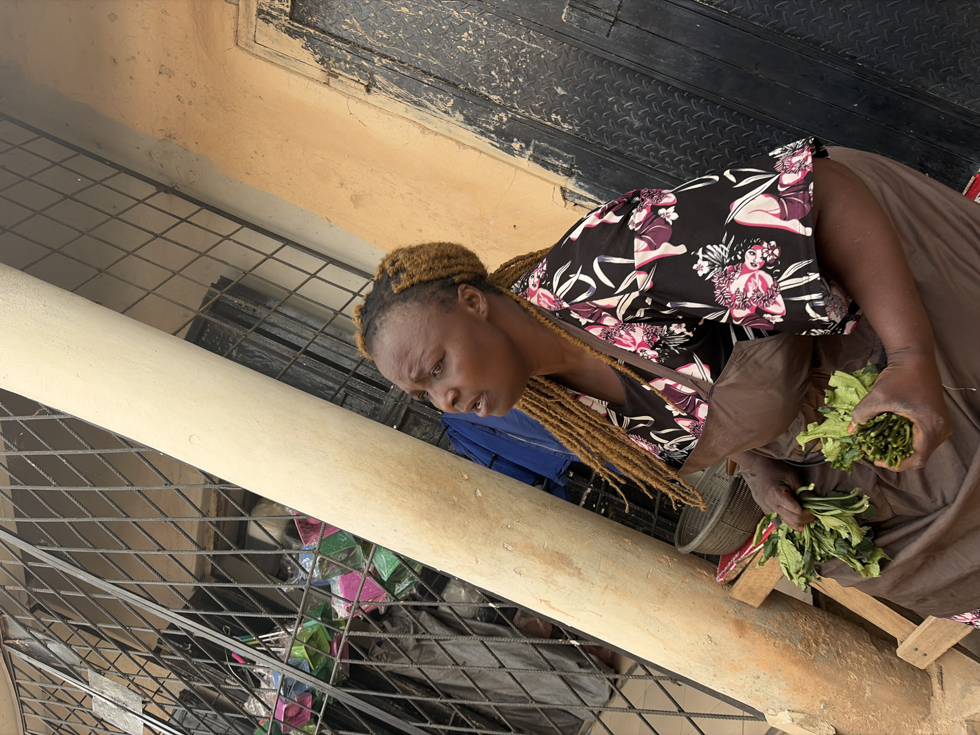 A woman is depicted in what appears to be an outdoor or semi-outdoor market setting in Jos, Nigeria. She has dark skin and her light brown, braided hair is styled upwards. She is wearing a black short-sleeved top adorned with white and pink floral patterns featuring figures of women, and a brown skirt or wrap. In her hands, she holds two bundles of fresh green leafy vegetables, suggesting she is a vendor or is purchasing produce. Her gaze is directed downwards towards the vegetables or something just out of frame, giving her a focused expression.
The background is characterized by a metal security grate or fencing that runs horizontally and diagonally across the lower and middle portions of the image. A thick, light-colored cylindrical object, possibly a pipe or pillar, crosses the frame diagonally. Behind the grate, colorful items wrapped in plastic are visible, hinting at other goods for sale. The ground beneath the woman is a rough mix of dirt and possibly concrete, while a dark, textured surface, possibly a metal door or wall, occupies the upper right background. The natural lighting indicates it is daytime. There is no visible text in the image.