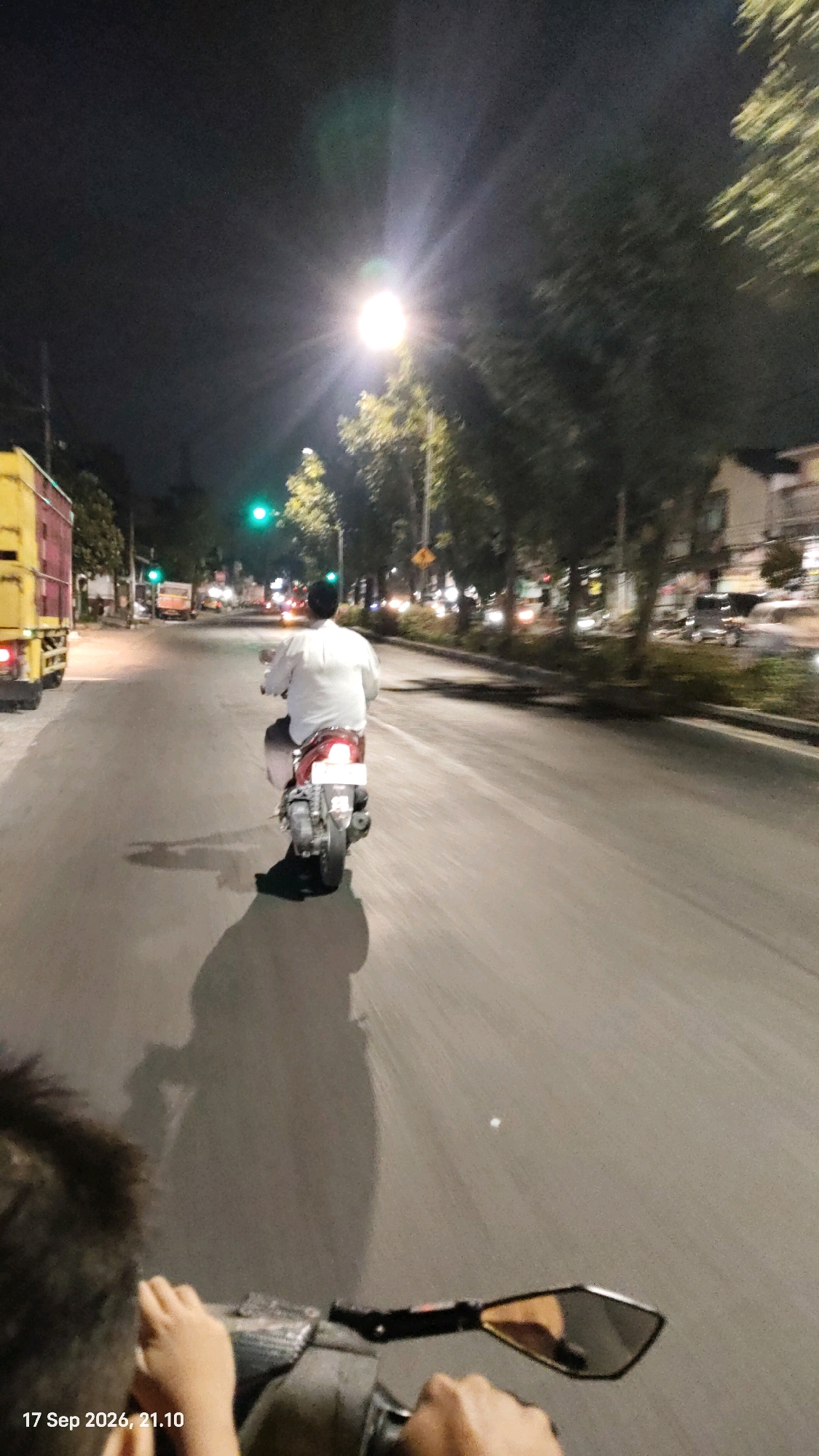 The image depicts a street scene at night in Surabaya, Indonesia. A person wearing a white shirt and a black cap is riding a red scooter on the right side of a paved road. The scooter has a white license plate. The rider's body is turned slightly to the left, and they are looking forward. In the foreground, a person's hands are visible, holding the handlebars of a scooter, suggesting the photo is taken from the perspective of another rider or passenger. A side mirror of this front scooter is also visible.
To the left of the road, a yellow and red truck is parked. Further down the road, there are streetlights, traffic lights (one green), trees lining the street, and buildings in the distance. The road surface is asphalt. The overall lighting suggests it is nighttime, with artificial lights from street lamps and vehicles illuminating the scene. The timestamp in the bottom left corner reads "17 Sep 2026, 21.10".