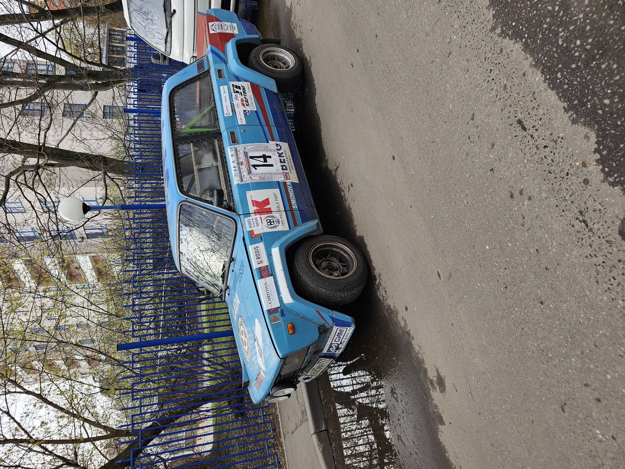 A light blue rally car with racing decals is parked on a street in an urban area. The car has racing number 14 on its side. The car is next to a dark blue fence. A portion of a white van is visible behind the rally car. The street is wet. Bare tree branches are visible in the background, along with a light pole and a building.