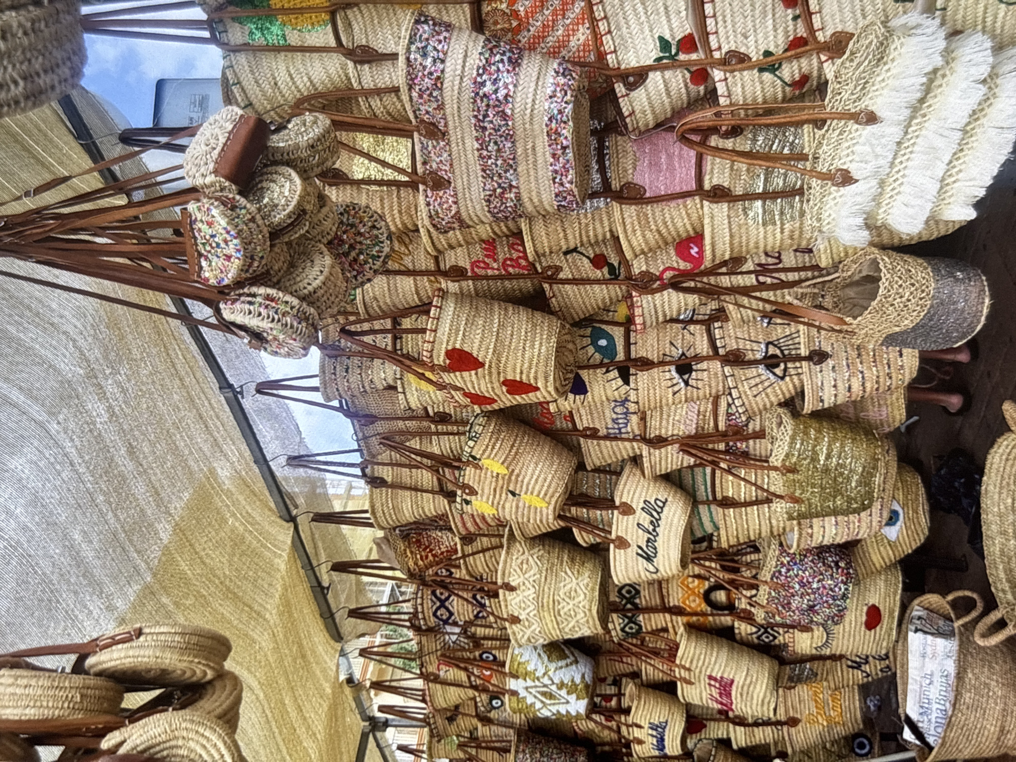 The image depicts a market stall filled with a large assortment of woven straw bags of various sizes and designs, hanging from a light-colored awning. Many of the bags feature colorful embellishments such as embroidered hearts, eyes, lips, cherries, and geometric patterns. Some bags are plain straw, while others have decorative bandings. There are also smaller, round coin purses and several larger tote bags with fringe detailing. In the lower left corner, a woven basket contains some folded papers. The overall impression is one of abundance and variety in handmade accessories. The location is identified as Orihuela, Spain, suggesting this is an outdoor market or a shop selling these items in that city. No people are clearly visible or interacting with the items.