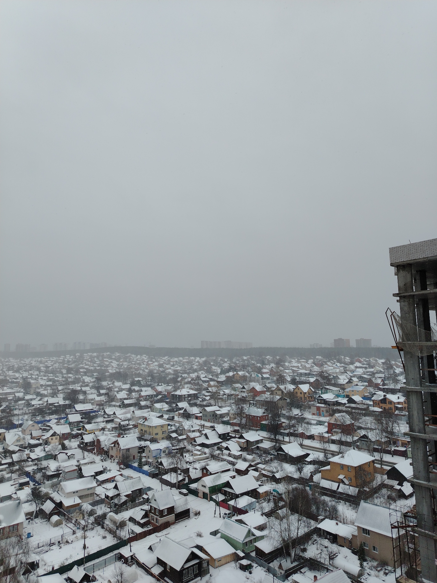 The image shows a snowy, overcast cityscape in Izhevsk, Russia. The roofs of numerous private houses are covered in snow. In the distance, beyond a sparse line of trees, are apartment buildings. To the right, there's a concrete construction site. No people are visible. The scene appears static, capturing a quiet winter day.