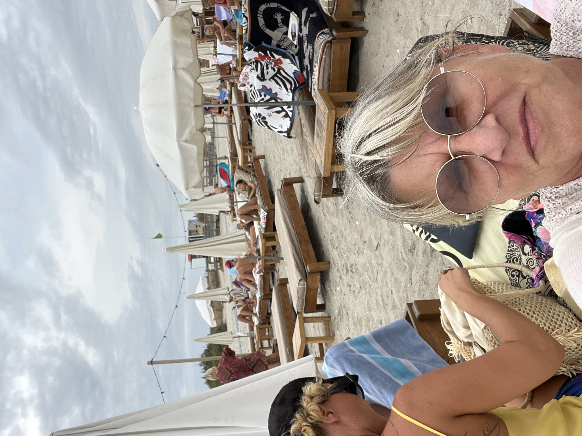The image depicts a beach scene in Frontignan, France, with several people relaxing on lounge chairs under umbrellas. The sky is overcast with clouds. In the foreground, a woman with blonde hair and round sunglasses is taking a selfie. To her left, another woman in a yellow top is visible from the torso down, holding a straw bag. In the background, various individuals are seen lounging, some appear to be reading or conversing. The beach is sandy, with rows of wooden lounge chairs and white umbrellas. A green flag is visible in the distance. The overall atmosphere suggests a relaxed day at the beach.