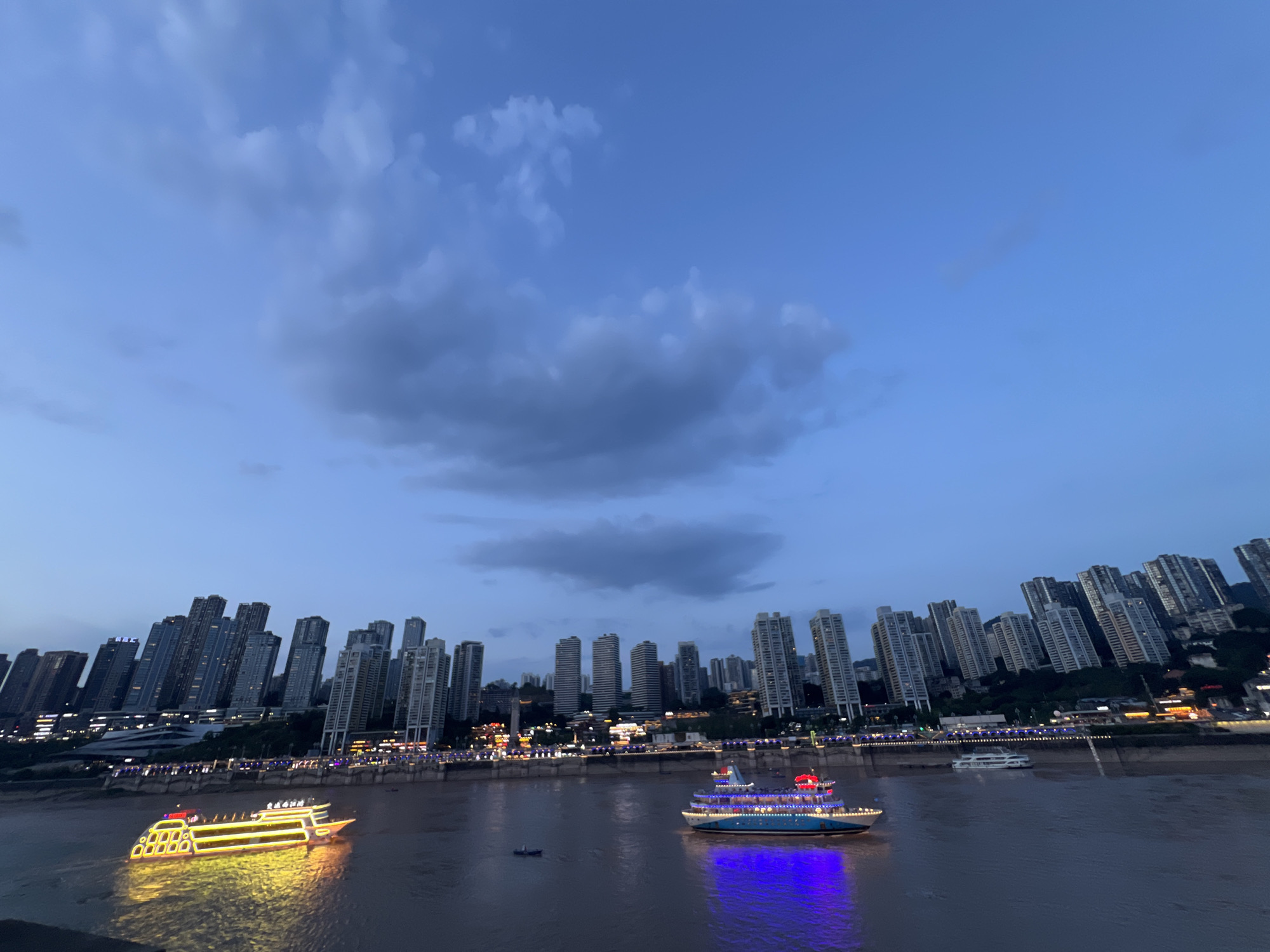 This image shows a cityscape at dusk in Yuzhong District, China. The sky is a deep blue with scattered clouds, and the city lights are beginning to illuminate the skyline. A wide river flows in the foreground, reflecting the lights from the city and two illuminated boats. The boats are of different sizes and designs, with one appearing to be a passenger cruise ship, adorned with bright yellow lights. The other boat, further in the distance, is smaller and appears to be a utility vessel, showing some blue and red lights. Along the riverbank, numerous tall buildings with modern architecture dominate the scene, their windows lit up. The buildings are arranged in a dense cluster, suggesting a high-density urban area. Some of the buildings are illuminated with colorful lights along their facades, adding to the vibrant nighttime ambiance. The overall impression is one of a bustling metropolis on the water, transitioning from day to night.