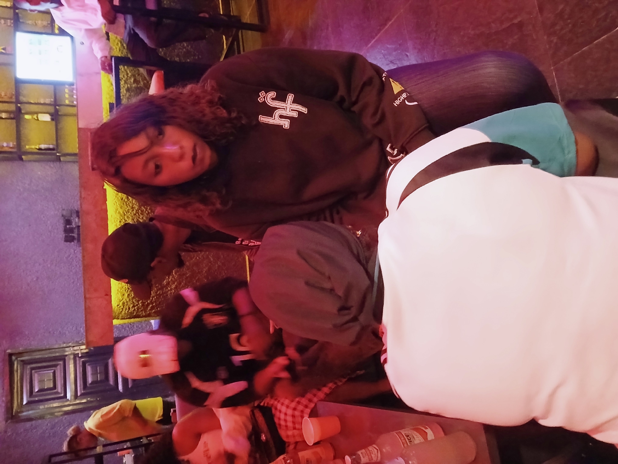A medium shot, taken at an angle, shows a group of people at a bar, likely on Rukuba Road, Nigeria. A woman with curly hair, wearing a black sweatshirt with white lettering, is looking at the camera. She is seated, partially obscured by someone wearing a white garment. In the background, other people are at a table with drinks, including what appear to be plastic cups and liquor bottles. The bar has shelves filled with bottles, and the lighting is dim, with a mix of purple and yellow hues. One person is wearing a cap, and the clothing styles suggest a casual setting.