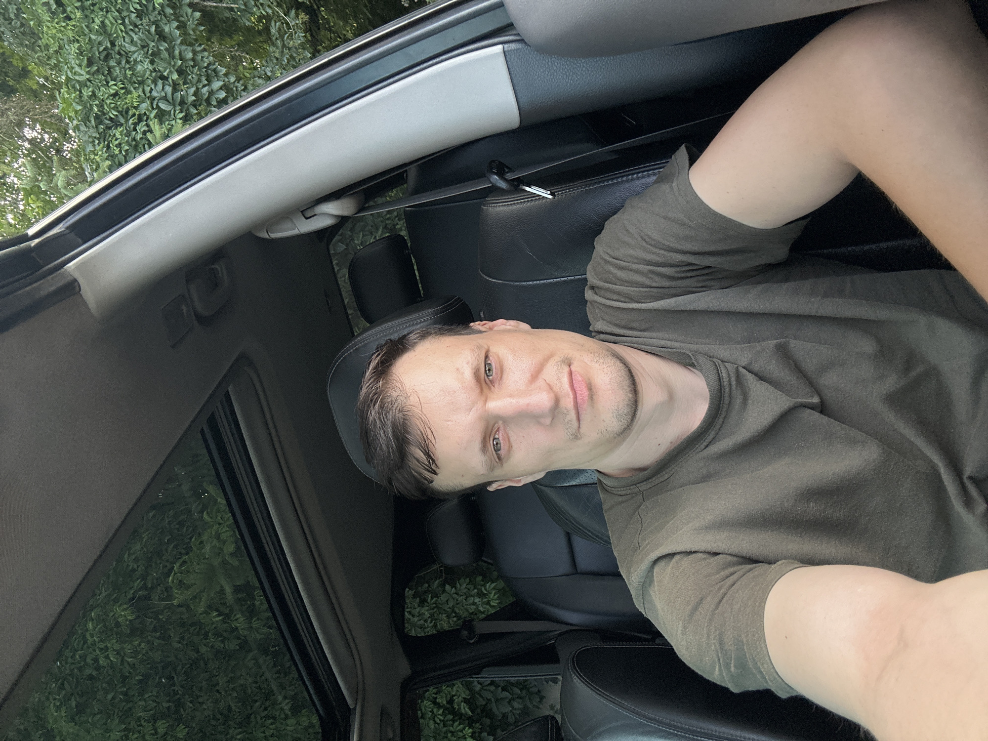 A man with short brown hair and light skin is sitting in the driver's seat of a car. He is wearing a dark green t-shirt. The car has black leather seats and a sunroof, which is open, revealing green foliage outside. The interior of the car appears to be dark. The man is looking directly at the camera with a neutral expression. The image was taken in Shatava, Ukraine.