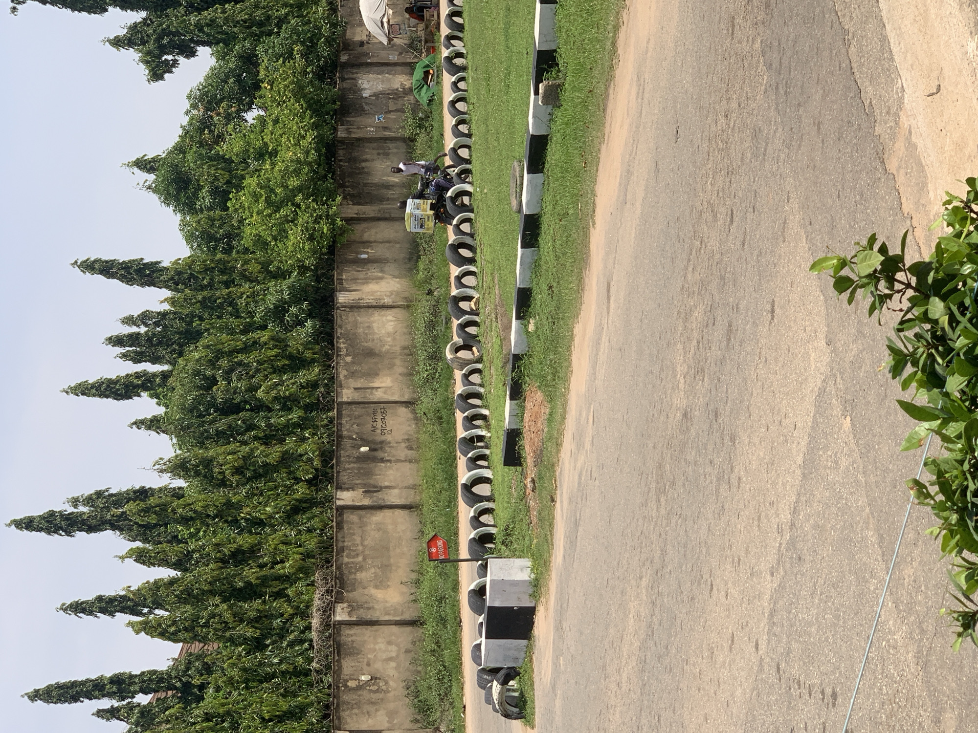 The image depicts a street scene in Abuja, Nigeria, featuring a roadway with a grass verge lined with stacked tires that serve as a barrier or track marker. Several tall, slender green trees are visible in the background, forming a dense line against a pale sky, with a concrete wall running behind them. On the grass verge, a red directional sign indicating "NO PARKING" is mounted on a pole. In the middle ground, two individuals are present: one person is riding a motorcycle with a yellow box attached to the back, and another person is walking nearby. Further in the background, a white tent structure is set up with people sitting beneath it, and a green tarp is visible. The foreground shows a paved road surface with some staining and a thin metal wire running across it. Lush green foliage is visible at the bottom of the frame.