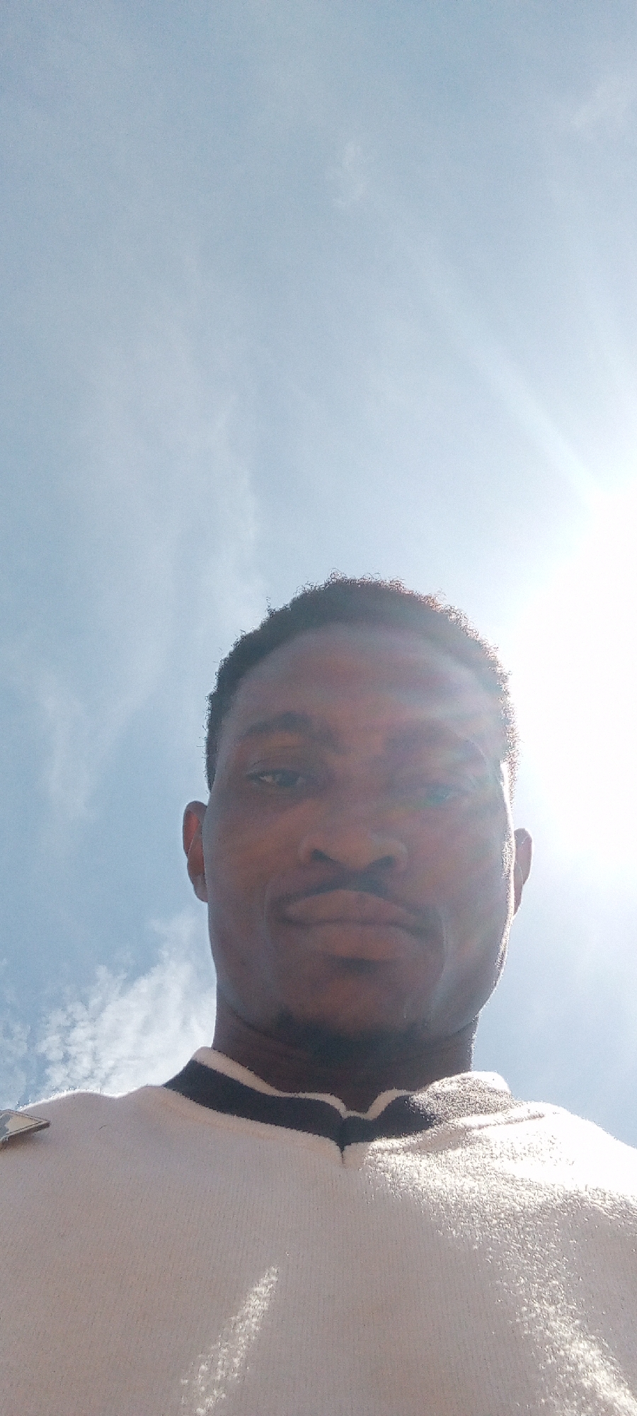 A low-angle selfie of a dark-skinned man is centered in the frame. He has short, dark hair and a mustache. He is wearing a white sweater with a dark stripe at the neck. The background shows a bright, partly cloudy sky with the sun shining strongly. The location is Adankolo, Nigeria.