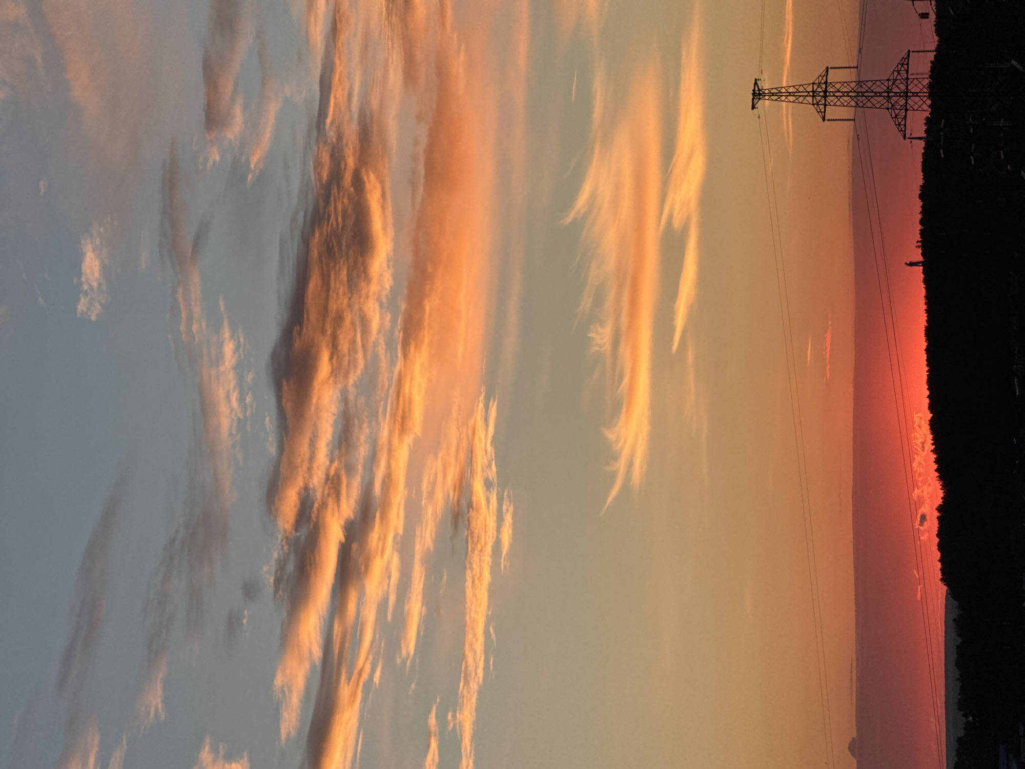 The image shows a sunset over the city of Chaikovsky, Russia. The sky is filled with vibrant orange and pink clouds, a result of the sun setting. In the foreground, there is a silhouette of a forest and a distant transmission tower. A single person is visible standing on a hilltop, silhouetted against the bright sky. The overall scene depicts a serene evening in a rural or suburban area.