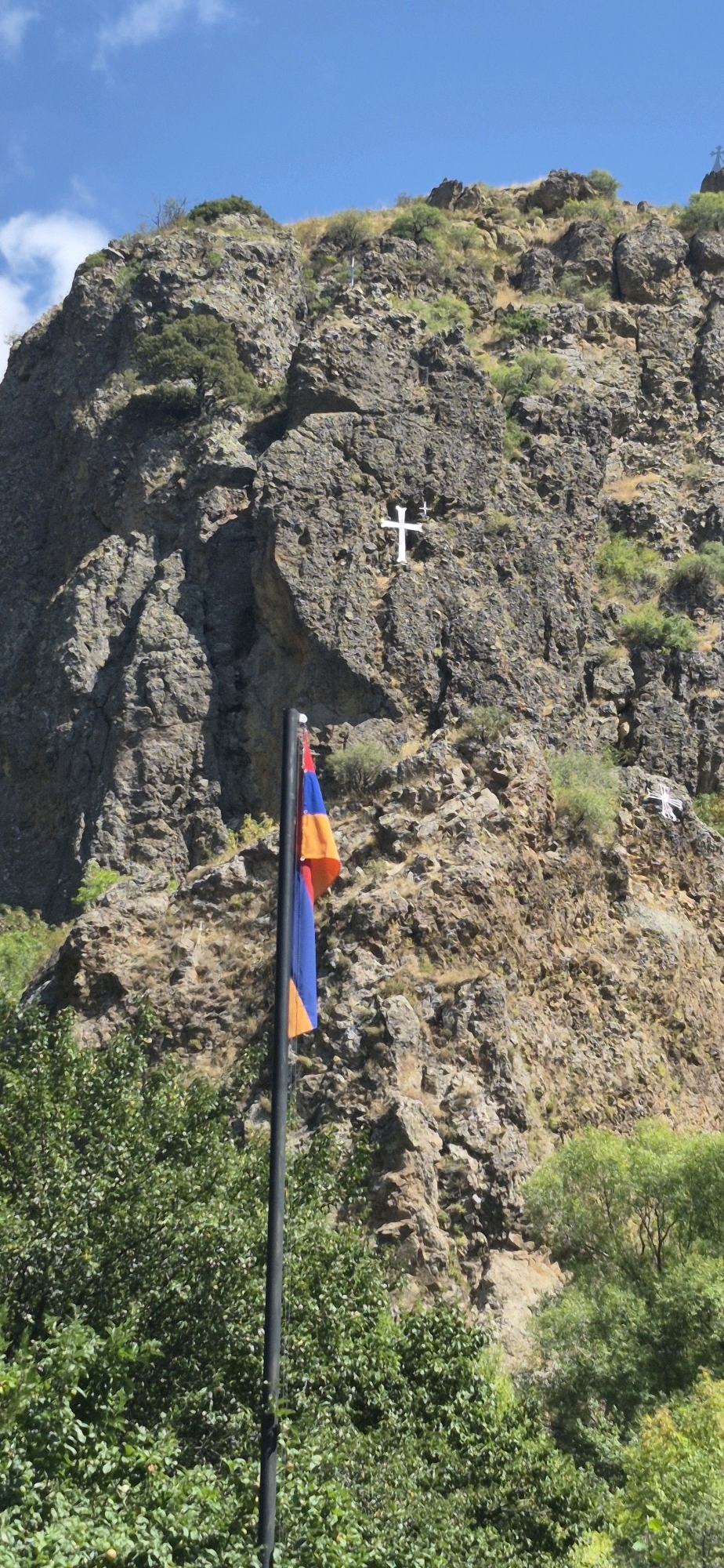 The image depicts a rocky mountainside under a bright blue sky with scattered clouds. A tall, dark flagpole stands in the foreground, bearing the red, blue, and orange striped flag of Armenia. Embedded in the rocky cliff face, to the right of the flagpole and slightly above the middle of the image, is a large, white cross. Further up and to the right, another smaller white cross is visible. Lush green trees and foliage partially obscure the base of the mountainside. The overall scene is likely captured in or near the village of Goght, Armenia, given the presence of the Armenian flag and the prominent religious symbol, which could be indicative of a significant historical or religious site in the region.