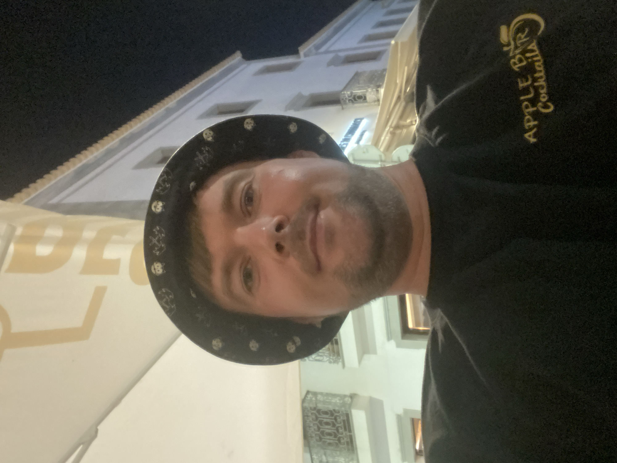 A man wearing a black hat with a pattern of small white skulls and a black t-shirt with a gold logo that reads "APPLE BAR Cocktails" is taking a selfie. The man appears to be outdoors at night, under a canopy. Behind him, a white building with several windows and balconies is visible. This building is consistent with the architecture found in the historic center of Tavira, Portugal.