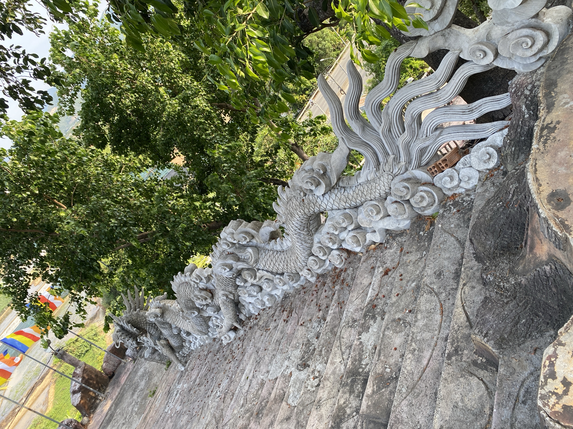 The image shows a stone staircase in Tay Nha Trang Ward, Vietnam, decorated with intricate dragon carvings that line the side of the stairs. The dragons are sculpted from grey stone, with detailed scales, claws, and flowing manes. They appear to be part of a temple or monument, given the presence of Buddhist flags in the background. The staircase itself is made of rough, weathered stone, showing signs of age and exposure to the elements. Lush green trees and foliage frame the scene, suggesting an outdoor, possibly mountainous or elevated location. The lighting indicates it is daytime, with sunlight casting shadows and highlighting the textures of the stone. There are no people visible in the image.