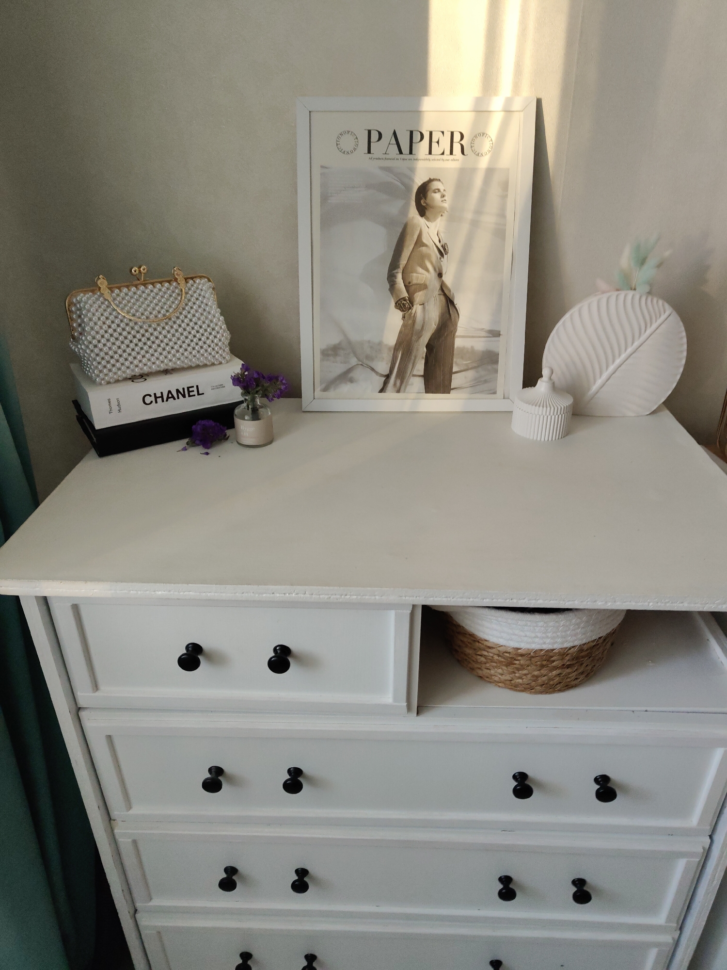The image shows a white dresser against a neutral-colored wall, possibly indoors. On top of the dresser, there's a decorative display with a white beaded purse resting on top of two books, one labeled "CHANEL." Next to the purse, there's a small vase with purple flowers. A framed image with the word "PAPER" printed on it is positioned near the center, next to a white decorative leaf-shaped object and a small, lidded container. The dresser has black knobs and multiple drawers. A partially visible basket with white and brown patterns sits in a drawer left slightly open. The room is illuminated by natural light, casting shadows on the wall behind the dresser. The location is Unknown, Unknown.