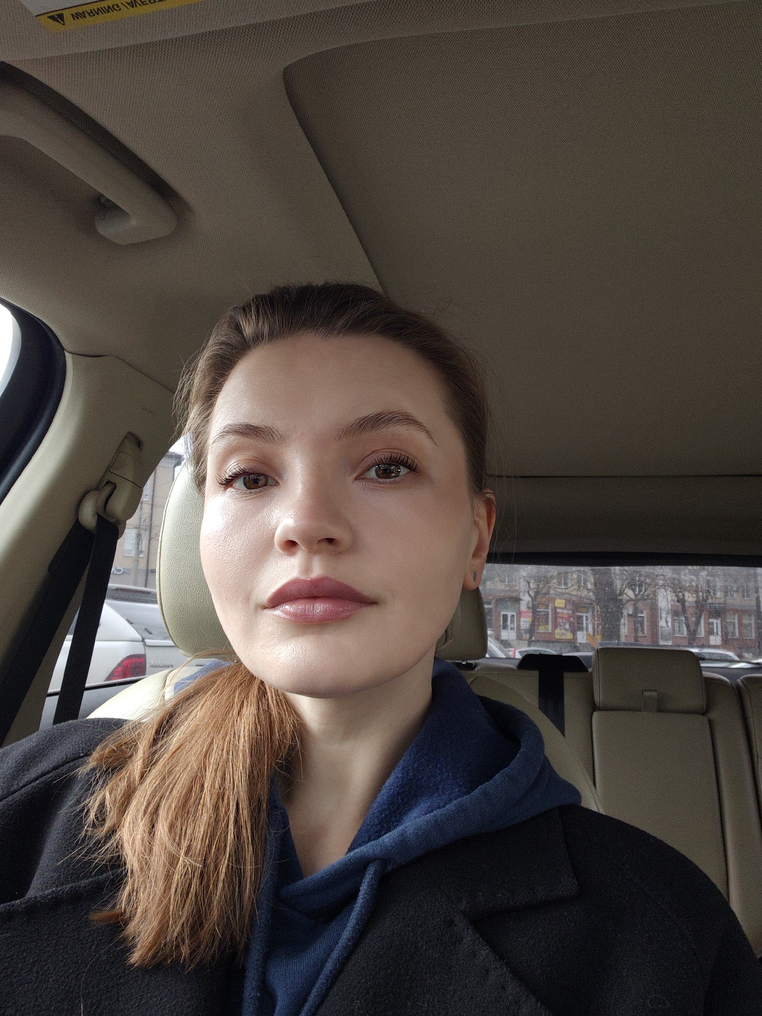 A woman is seated in the driver's seat of a car, facing the camera. She has fair skin, brown eyes, and light brown hair pulled back in a ponytail. She is wearing a dark coat over a blue hoodie. Outside the car window are buildings, other cars, and trees. It appears to be daytime and the weather is overcast.