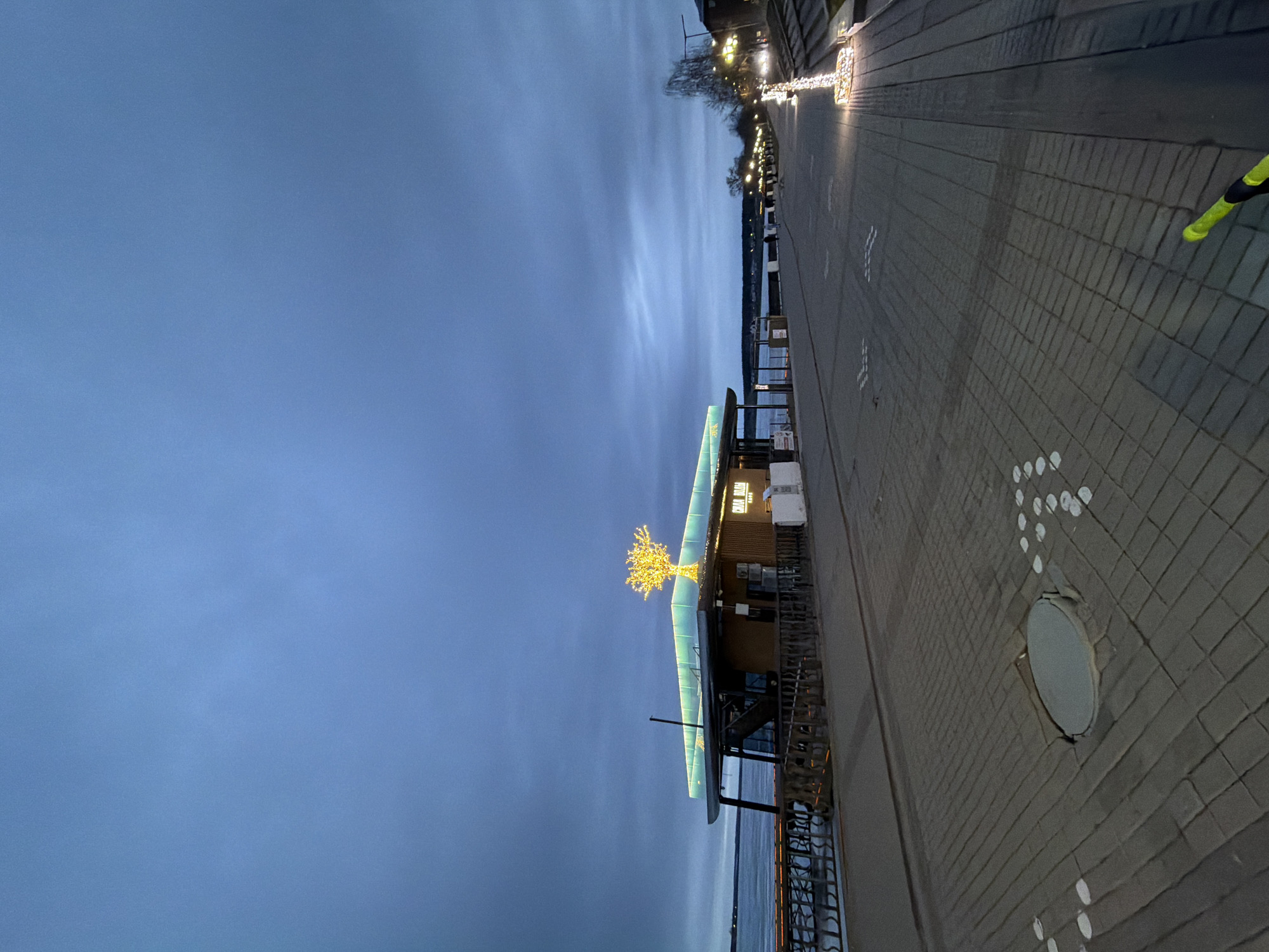 The image shows a building and adjacent paved walkway along a body of water in Votkinsk, Russia. The building has a blue-green roof and a small tree with yellow lights on the top. It appears to be a cafe, with a sign visible that says "CEDAR HOUSE CAFE." To the right of the building is a paved walkway with white markings and a circular metal access cover. Street lights illuminate the walkway. The sky is blue, tinged with dusk. A sliver of water is visible at the bottom.