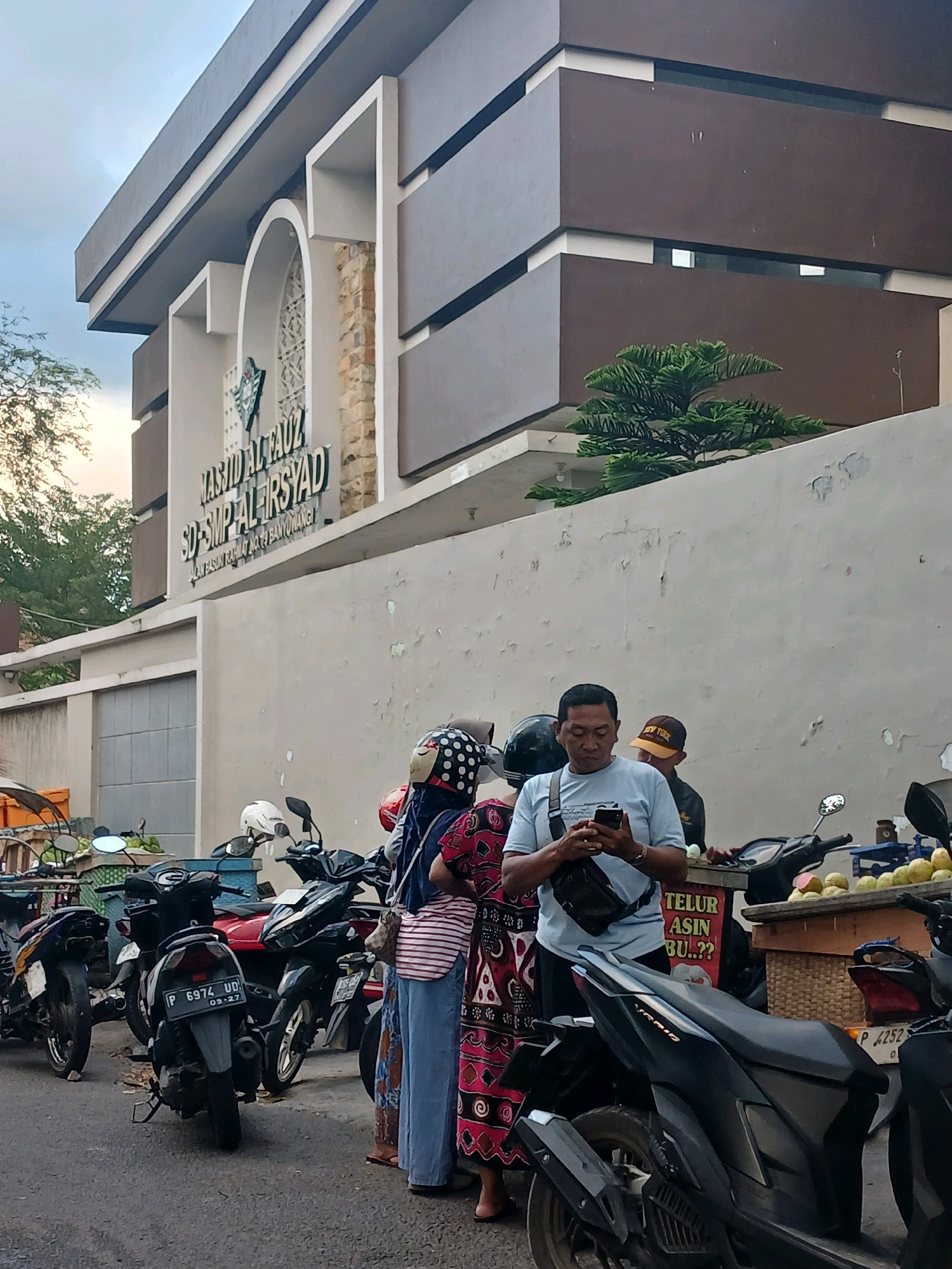The image depicts a street scene in front of a building identified by a sign as "MASJID ALFAUZI SD-SMP AL IRSYAD". The architecture of the building features a modern design with brown and gray rectangular elements and arched windows. In the foreground, several motorcycles are parked. Three individuals are visible near the motorcycles: a man in a light blue t-shirt is looking at his phone, a woman in a red and black patterned dress is partially obscured, and another person wearing a helmet is standing behind the man. A fruit stand with green fruits and a sign that says "TELUR ASIN" (salted eggs) is also present. The setting appears to be a public area, likely a street or sidewalk, in Banyuwangi, Indonesia, given the license plate format of the motorcycle "P 6974 UD". The sky is overcast, suggesting it might be late afternoon or early morning.