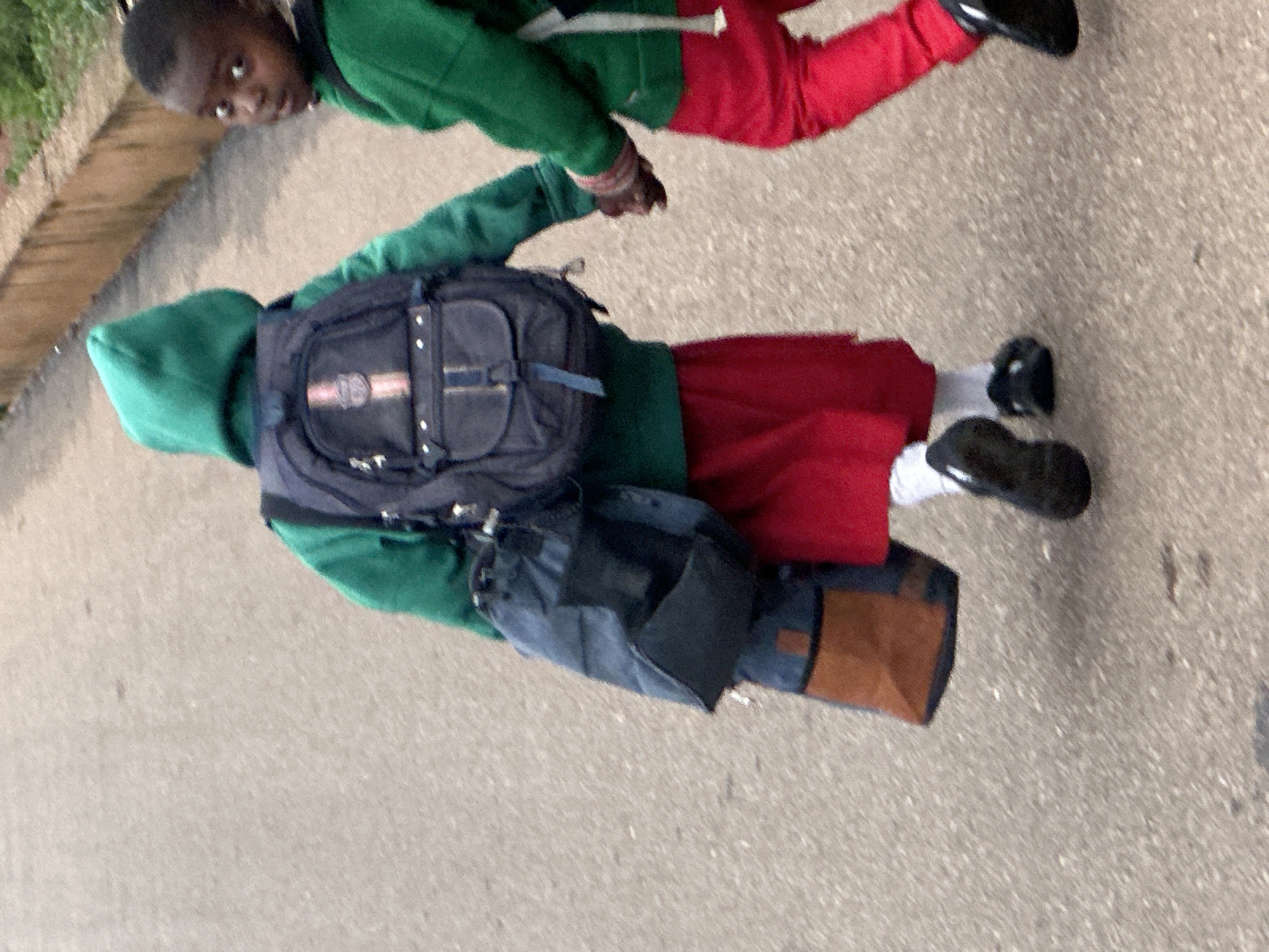 Two children, likely of Nigerian ethnicity, are walking on a paved surface. The child in the foreground, facing away from the camera, wears a green hooded jacket, red shorts, white socks, and black shoes. They are carrying a blue and black backpack. To the right, another child, facing the camera, wears a green jacket and red pants. The background features a low concrete wall with some vegetation. The overall scene suggests a street or school environment in Gura Top, Nigeria.