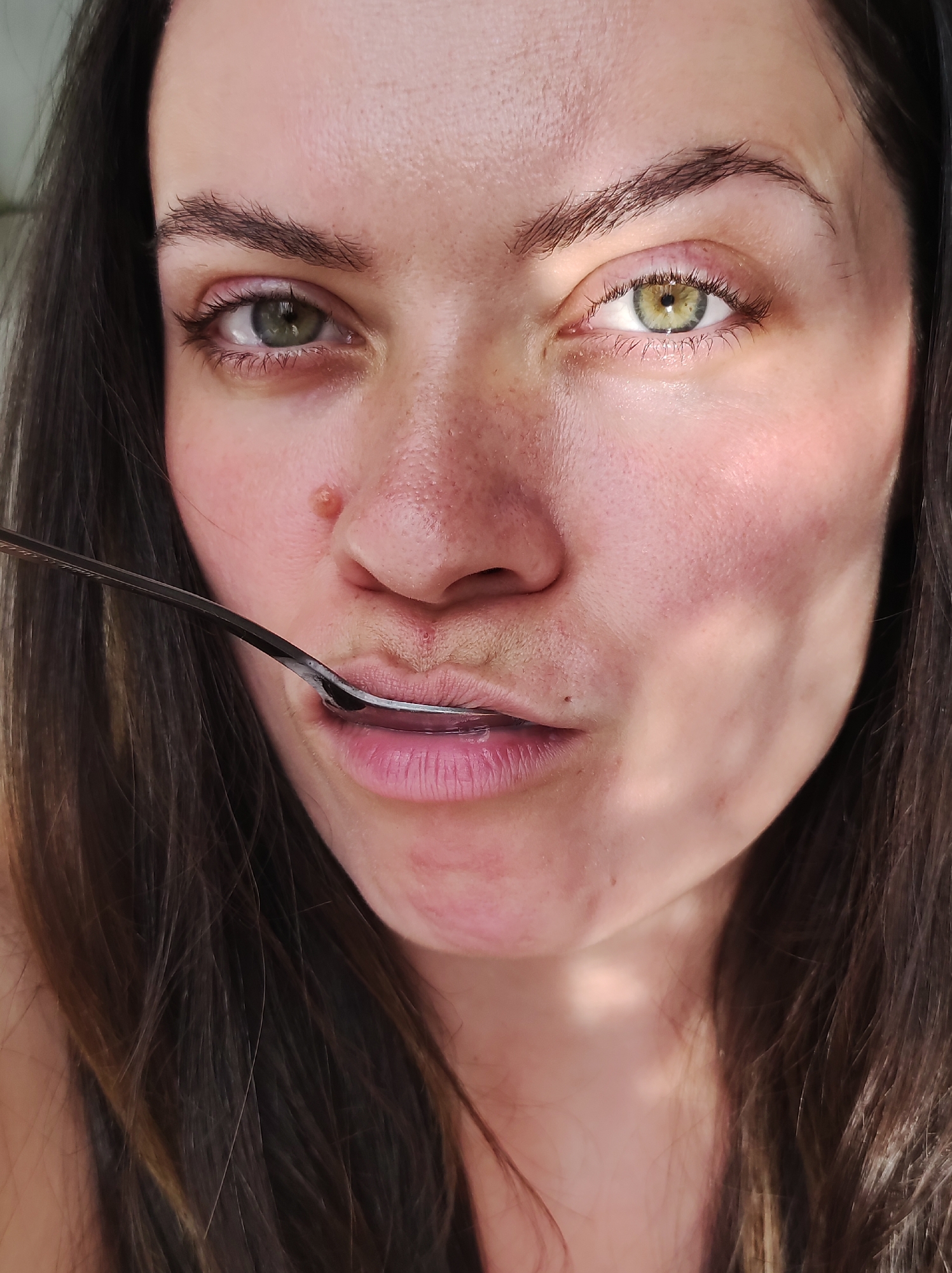 A close-up shot shows a woman's face with fair skin and dark hair. She holds a silver spoon horizontally across her lower lip. Her eyes are hazel-green, and she has a mole on her nose. The lighting appears to be natural and diffused. The city or location cannot be determined from the provided image.