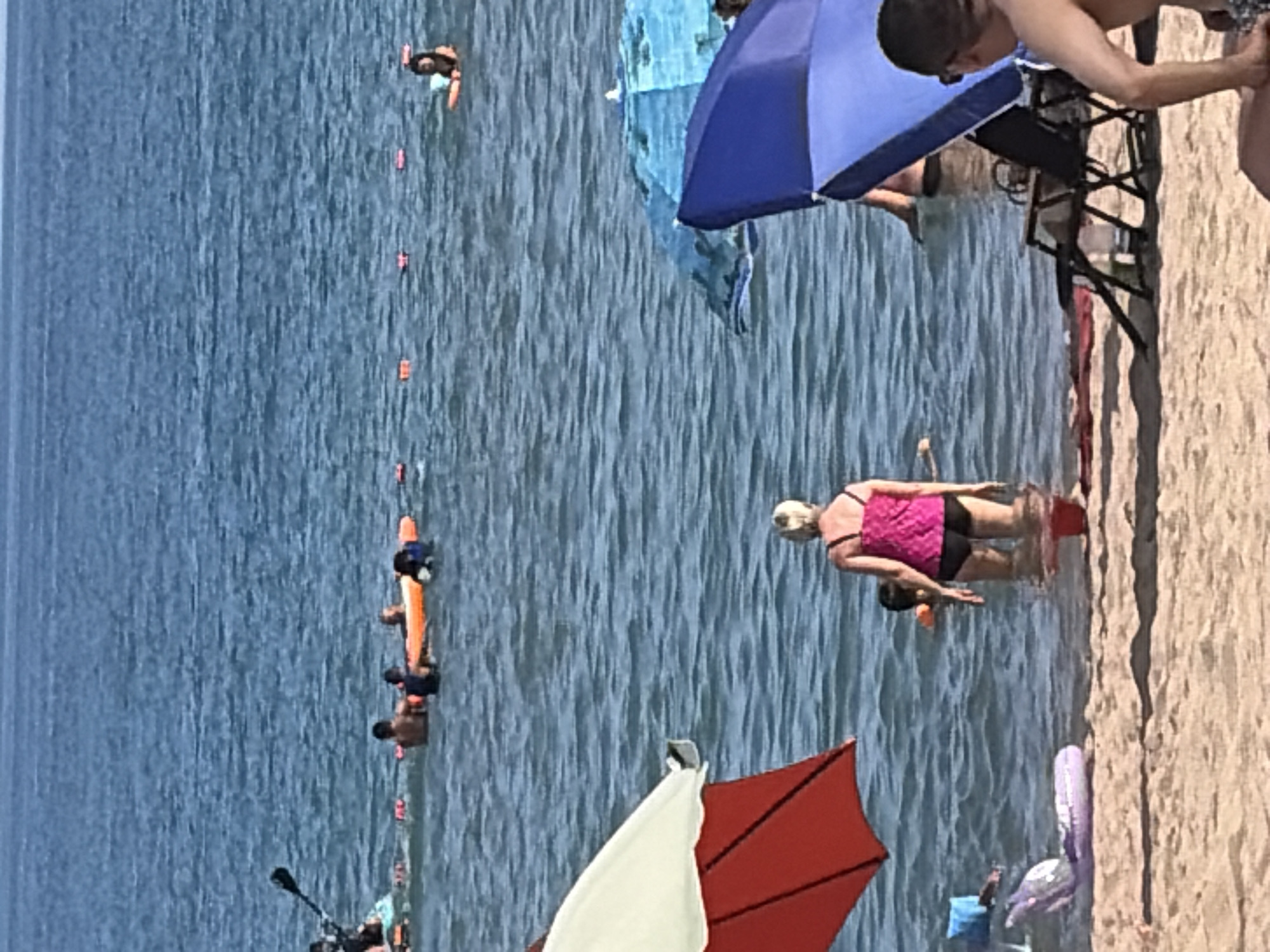 In an outdoor, daytime scene at a beach, several people are enjoying the water and the sand. The water is blue and appears calm, with small waves. In the foreground, a beach umbrella with a red and white canopy is partially visible on the left. Further out in the water, a woman in a pink swimsuit and black shorts stands with her back to the camera, facing the sea. To her right, a blue umbrella is open, and a person wearing sunglasses is seated in a beach chair next to it. In the middle ground, a group of people are in the water, some swimming and others relaxing on inflatable objects. One person is on a paddleboard in the distance on the left. The beach itself is sandy, with some red fabric and a purple inflatable lying on the shore in the foreground.