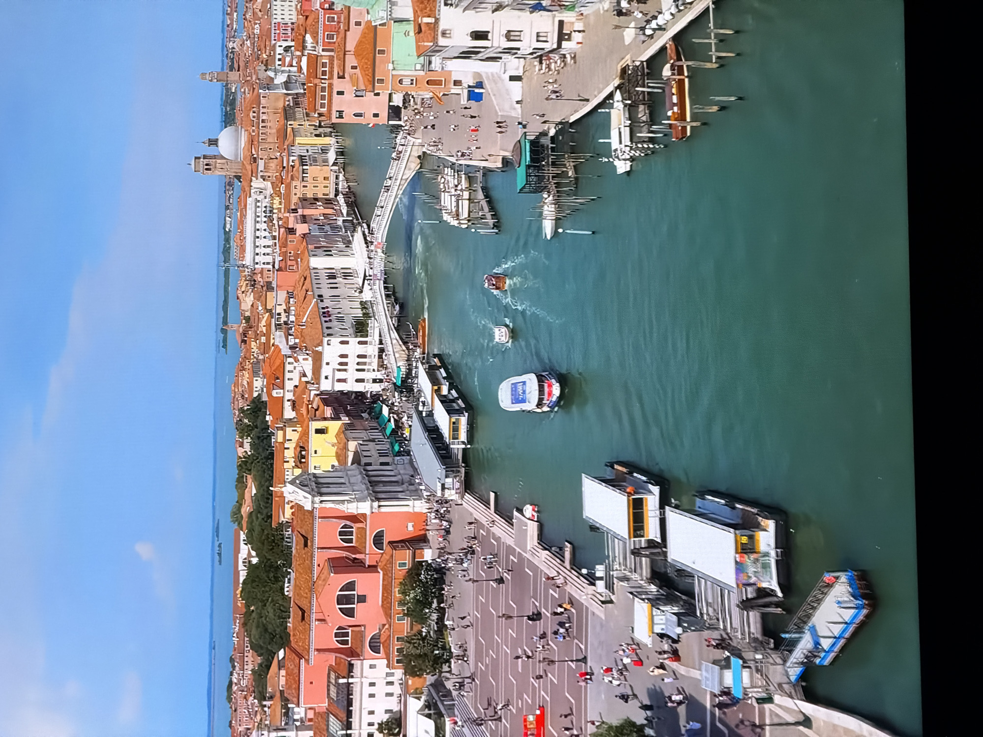 The image depicts a wide-angle, high-angle view of a canal in Venice, Italy, under a clear blue sky. The canal is filled with various boats, including passenger ferries, smaller motorboats, and what appears to be a gondola. The water is a deep turquoise color. Along the canal banks are numerous buildings with terracotta roofs, typical of Venetian architecture. Some buildings are brightly colored, while others are white or cream. Pedestrian walkways line the canal, with many people walking and gathered. Several bridges are visible crossing the canal in the distance. The scene is bustling with activity, suggesting a tourist area. The question asks to include the location of Chirchik, Uzbekistan, however, this city is not depicted in the image.