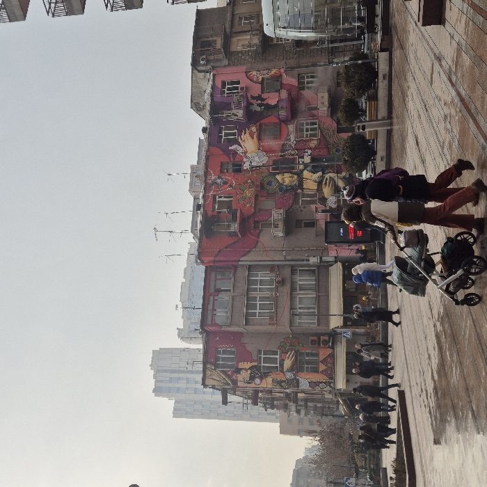 The scene depicts a public urban square on a cloudy day in Yerevan, Armenia, specifically on Aram Street, near the intersection with Abovyan Street in the Kentron district. The ground pavement is damp. A prominent multi-story building occupies the midground, featuring a large, vibrant mural across its entire facade. The mural, rendered in shades of pink, purple, orange, and cream, depicts stylized female figures, faces, hands, and intricate patterns. Modern high-rise buildings are visible in the distant background.
In the immediate foreground, two individuals walk from right to left. The leading person, wearing a light-colored jacket and rust-toned pants, pushes a dark-colored baby stroller. To their left, a second person, dressed in a dark jacket and similar rust-toned pants, walks alongside, carrying a purple backpack. In the midground, numerous pedestrians, clad in winter clothing, traverse the paved square in various directions. Street furniture, including planted shrubs and a modern glass-enclosed structure, is visible on the right. An illuminated digital display board with red text is situated near the mural building. Antennas and satellite dishes are present on the rooftops of visible buildings.