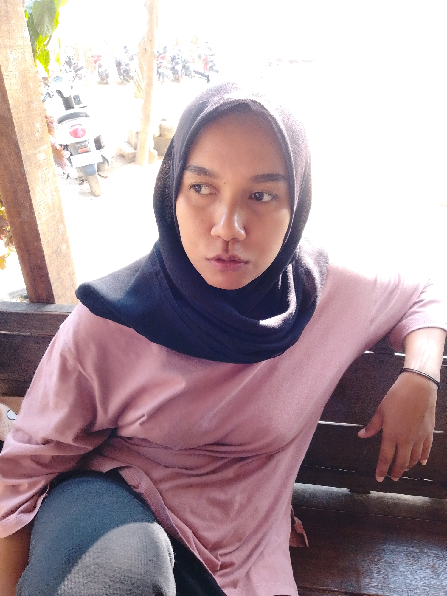 A medium shot shows a woman wearing a black hijab and a pale pink long-sleeved shirt. She is seated on a wooden bench, leaning her left arm on the back of the bench, with her right arm resting on her lap. The woman's gaze is directed to her left, away from the camera. Her skin tone is medium brown. A thin black bracelet adorns her right wrist. The background is out of focus, but appears to be an outdoor setting with motorcycles and possibly trees, suggesting a location in a village or a less urbanized area of Cikal, Indonesia. Sunlight is filtering in from the right, creating bright highlights and casting some shadows. The overall impression is that of a casual, personal moment captured in natural light.
